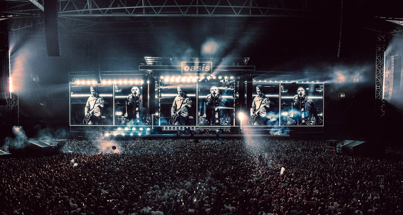 Concert stage with the band Oasis performing, large screens displaying band members, bright stage lights, and a massive crowd of fans.