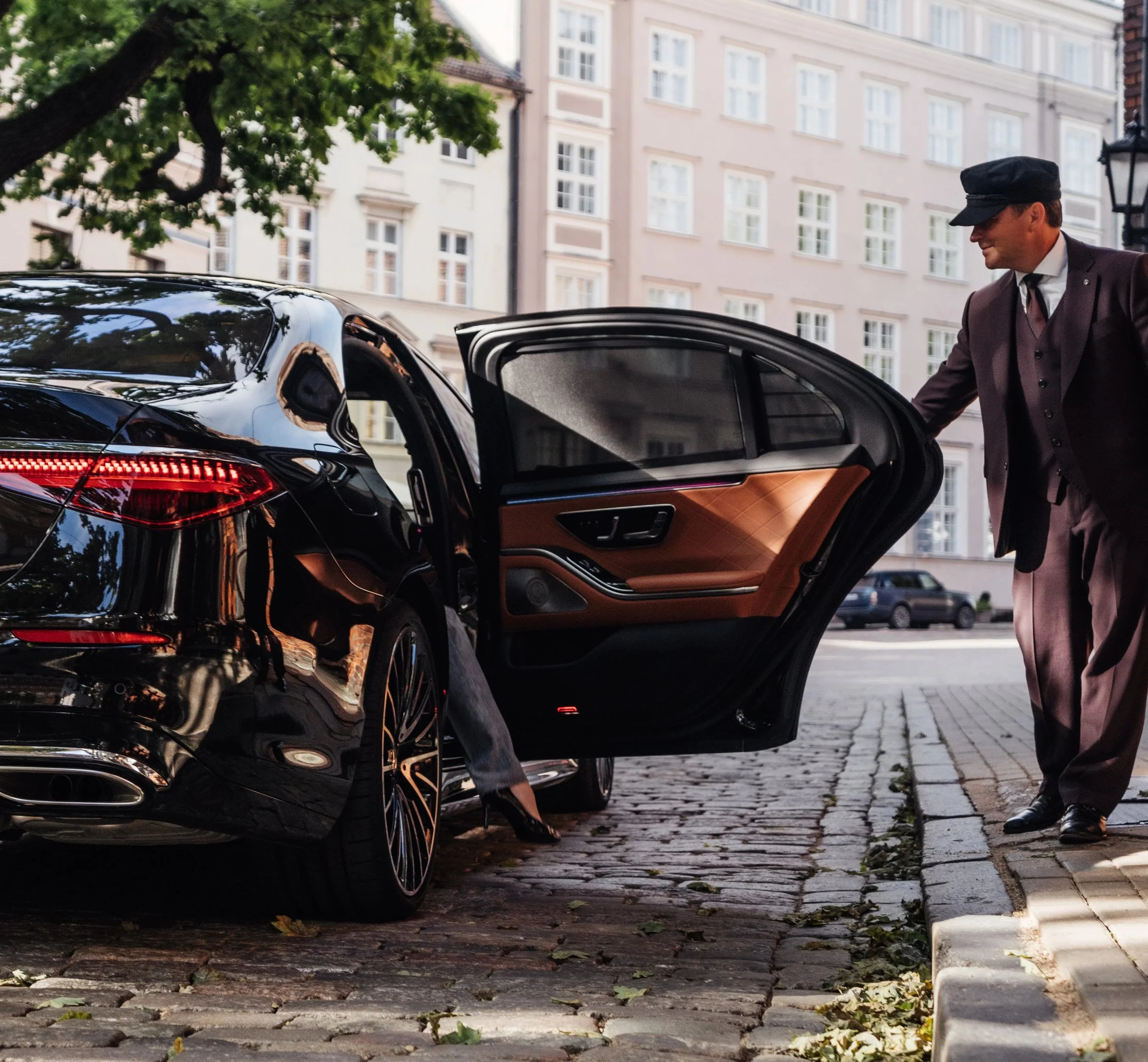 A chauffeur opening the rear door of a black luxury car for a passenger on a city street with cobblestone pavement, multi-story residential buildings, and trees in the background.