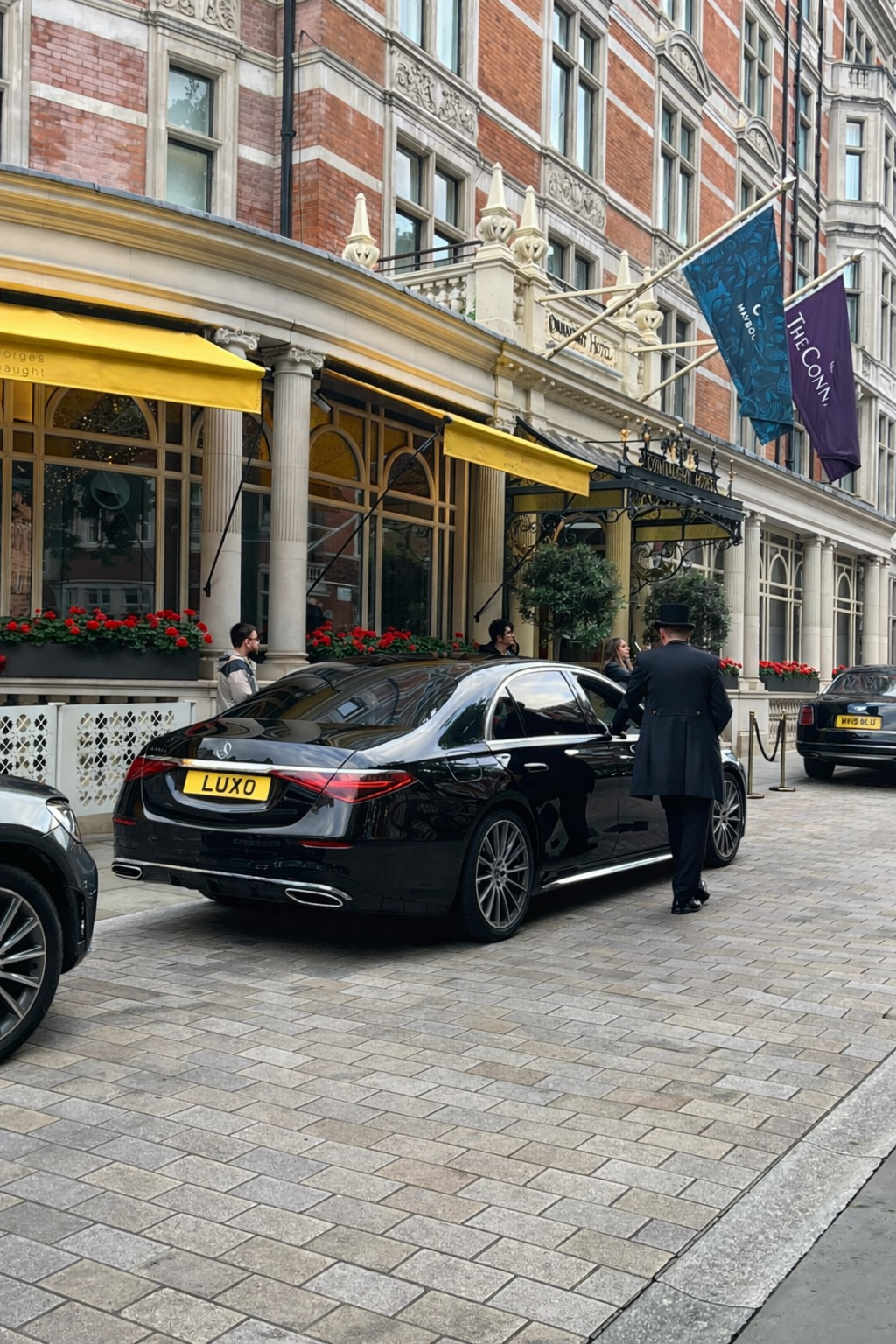 Luxury black car parked in front of a hotel entrance, with a man dressed in a formal coat and hat standing nearby. The hotel has decorative architecture with flags and red flower arrangements.