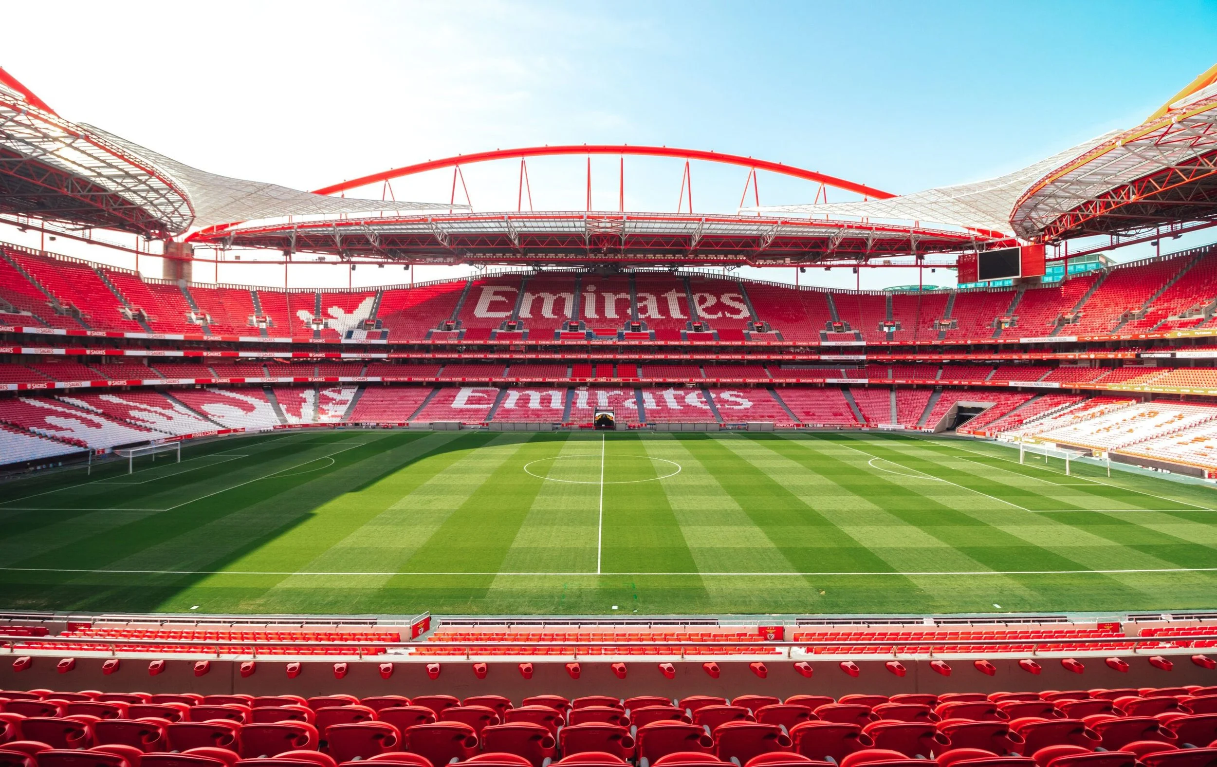 Empty soccer stadium with red seats and green field, 'Emirates' logo on the stands, clear blue sky.