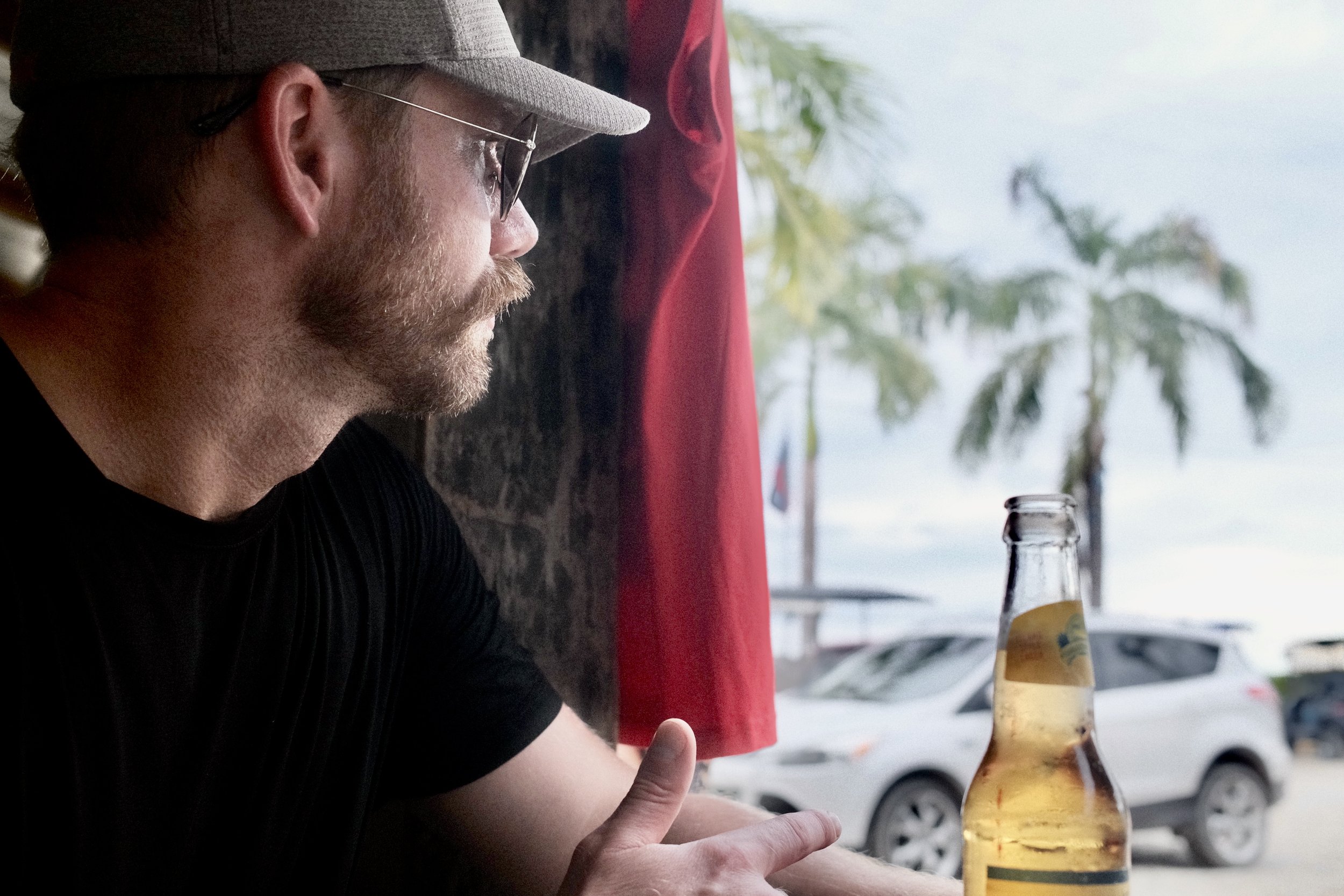 A man with glasses and a beard sitting at a table in a cafe, looking out the window at palm trees and parked cars, with a bottle of beer in front of him.
