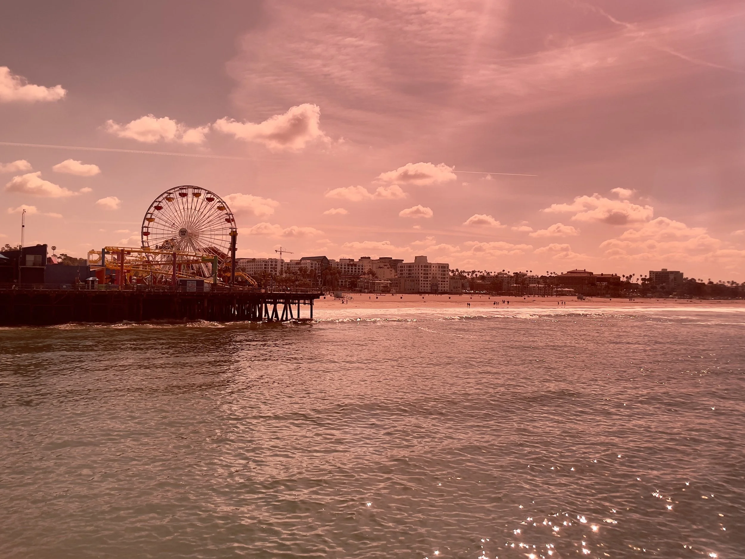 A beach scene with a pier and a large Ferris wheel, against a pink sky with clouds and distant buildings in Santa Monica, CA