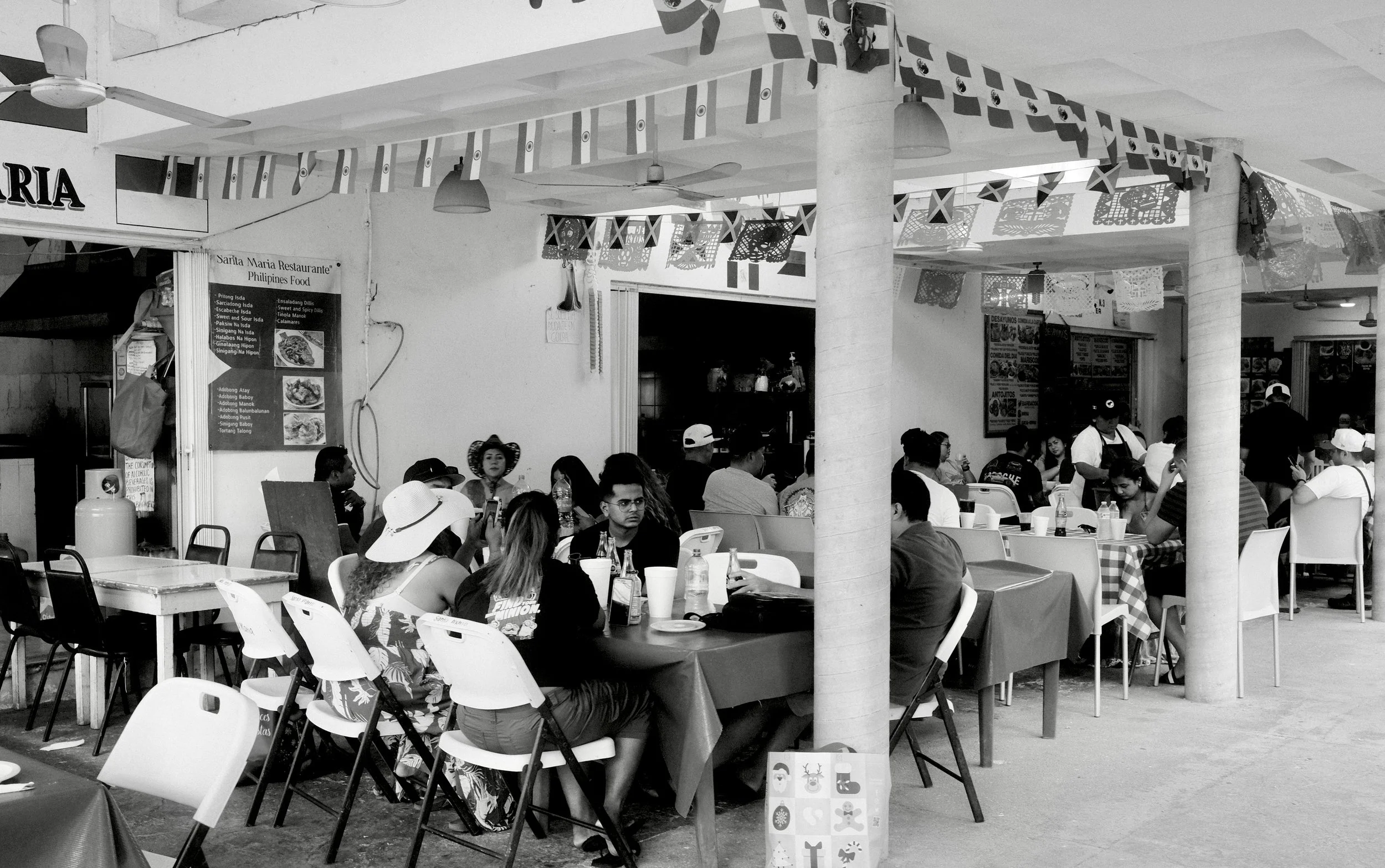 People dining in an outdoor restaurant decorated with small flags and banners, with tables covered in tablecloths and an assortment of bottles and cups on the tables.