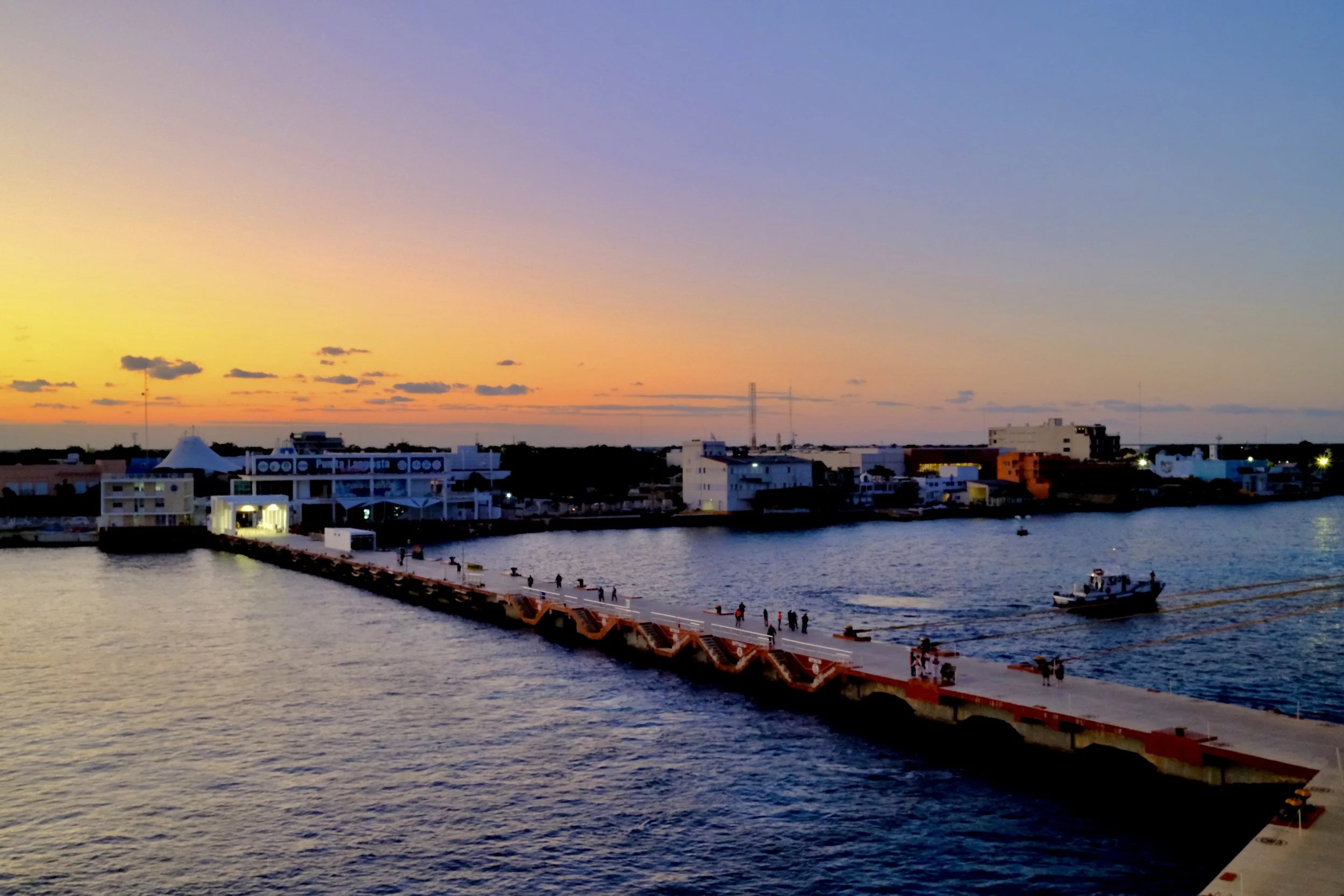 Sunset over a coastal city with a pier, boats on the water, and buildings along the shoreline.