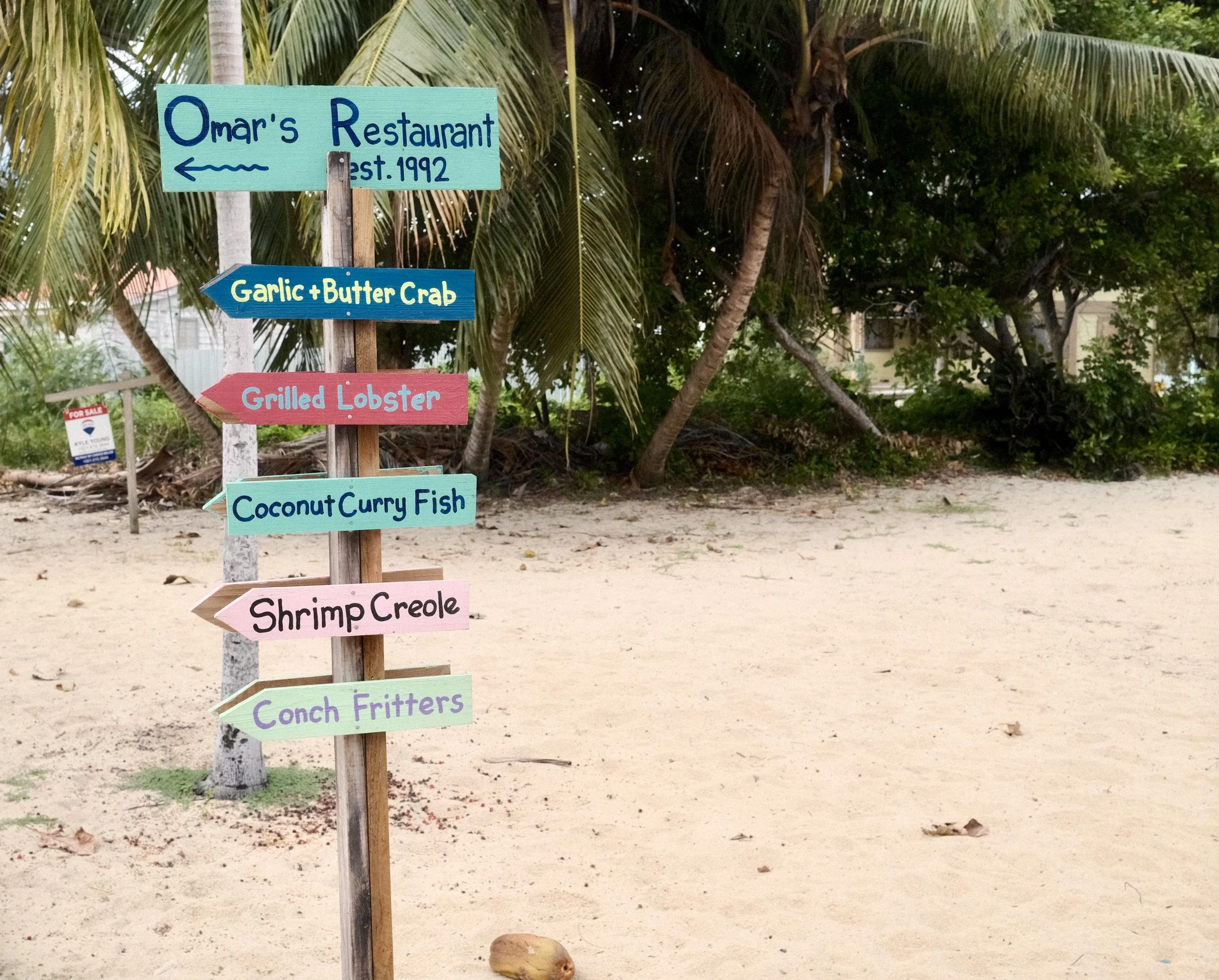 Colorful directional signs on a beach showing different food options, backed by trees and sand.