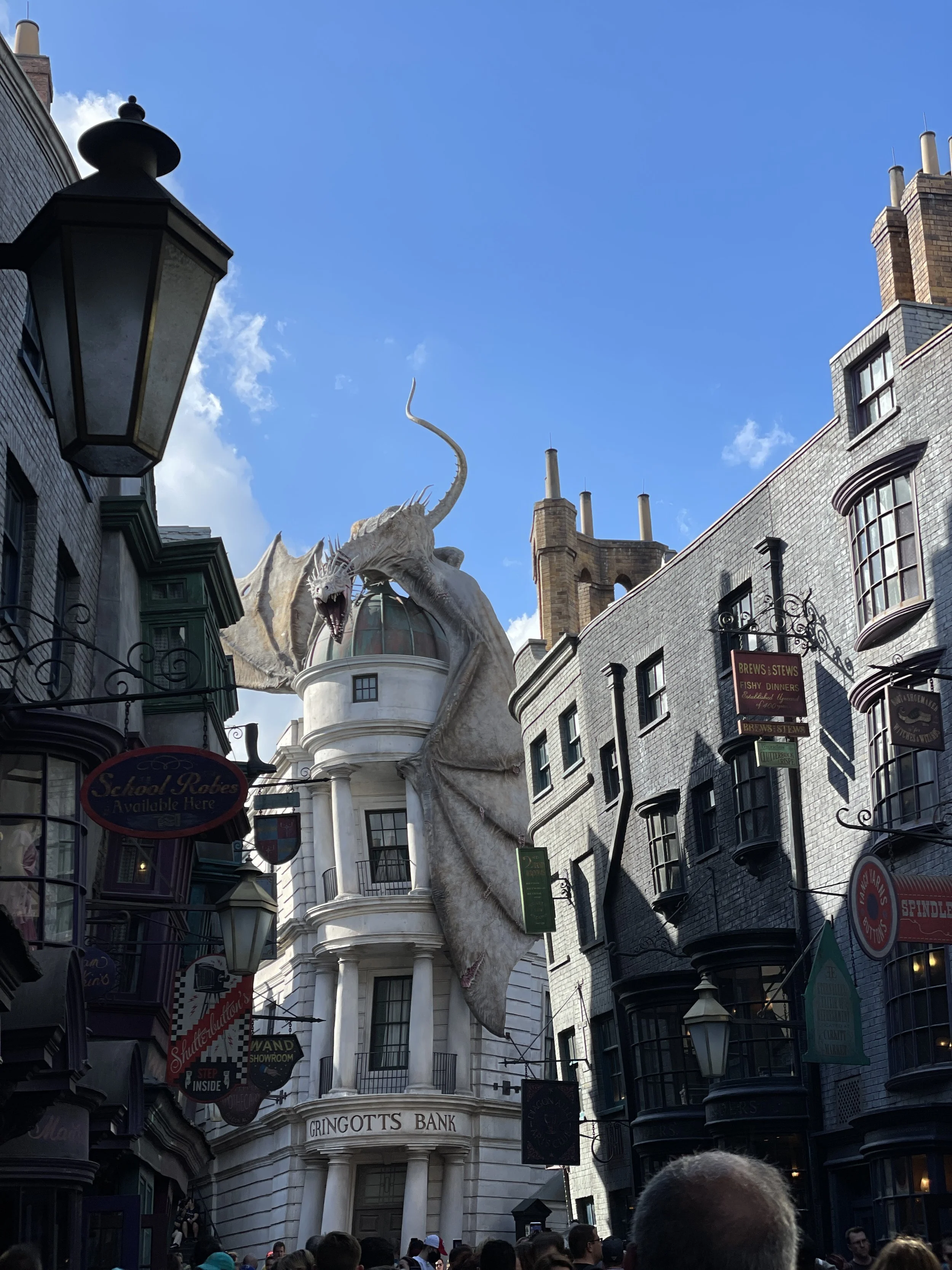 A large, detailed dragon sculpture perched over a building with the sign 'Gringotts Bank' in a busy street with various store signs and a crowd of people.