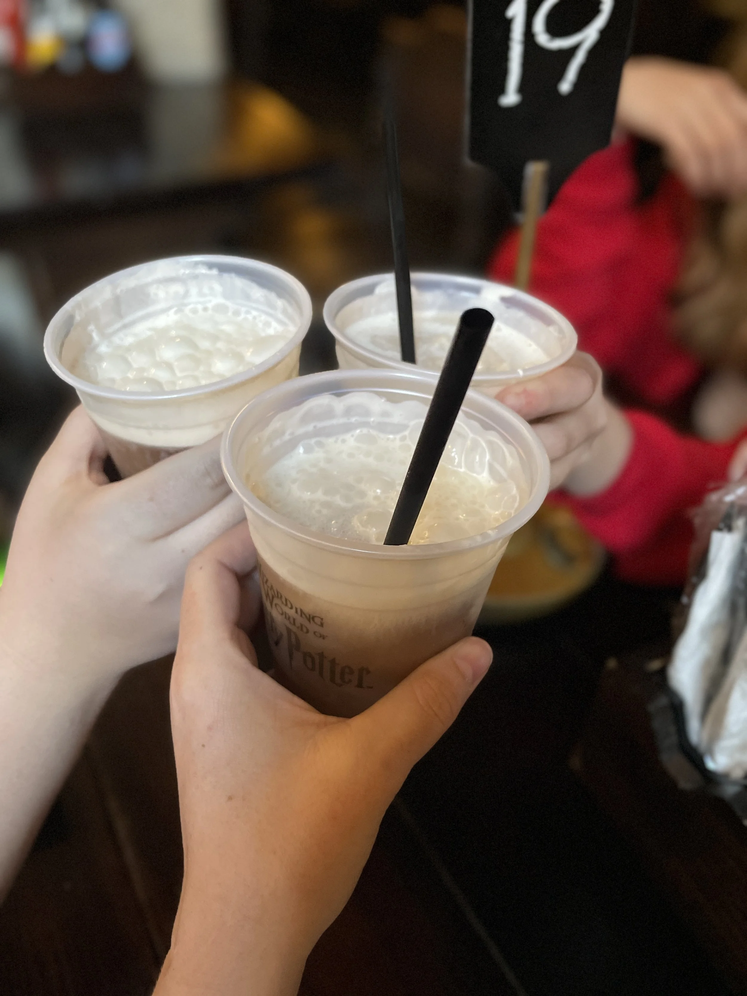 Hands holding three cups of coffee with whipped cream, toasting in a social setting, with a table and a person in a red shirt visible in the background.