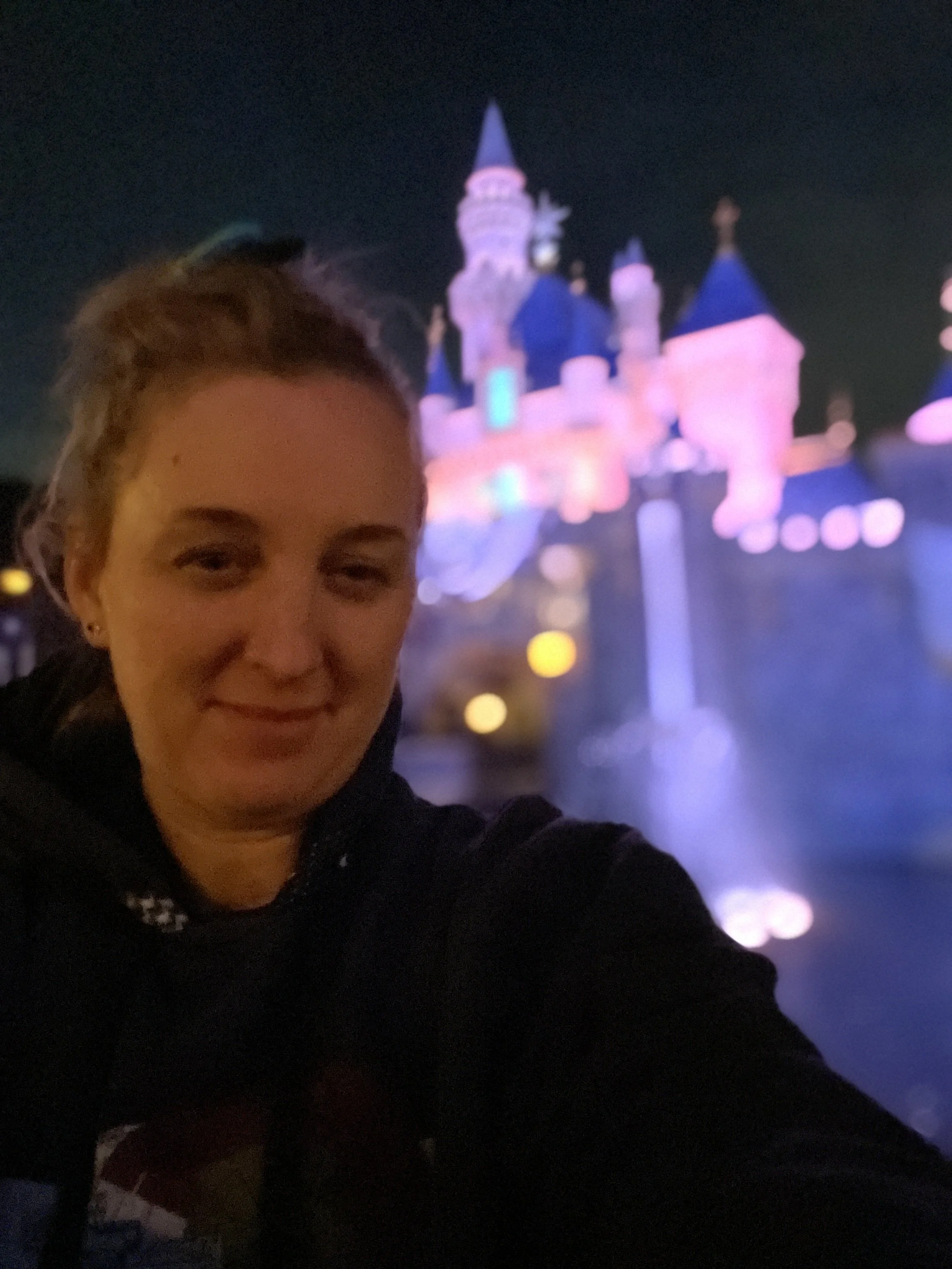 A woman taking a selfie at night with a Disney castle illuminated in pink, purple, and blue lights in the background.