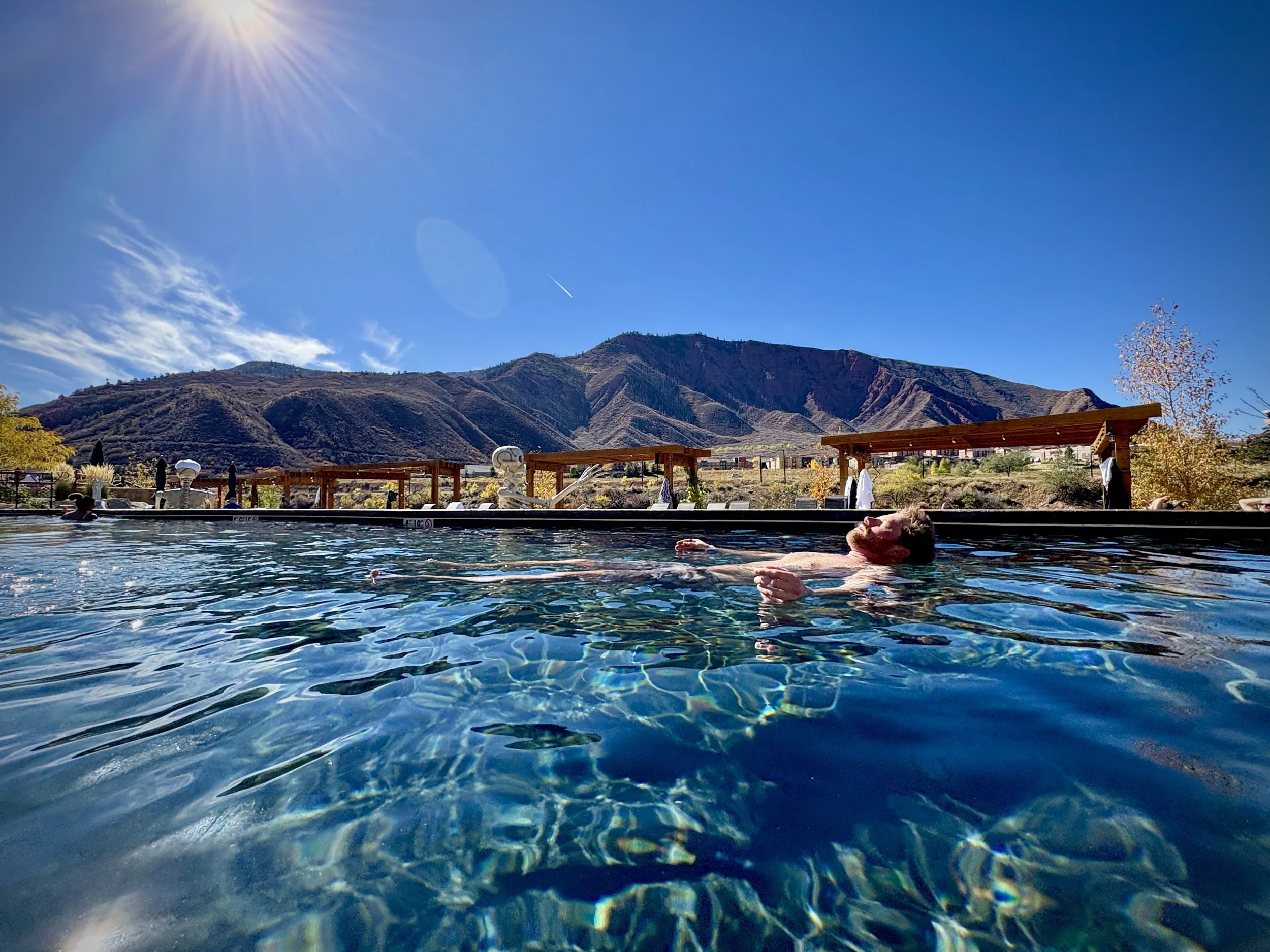 Man relaxing in an outdoor swimming pool with mountains in the background on a sunny day.