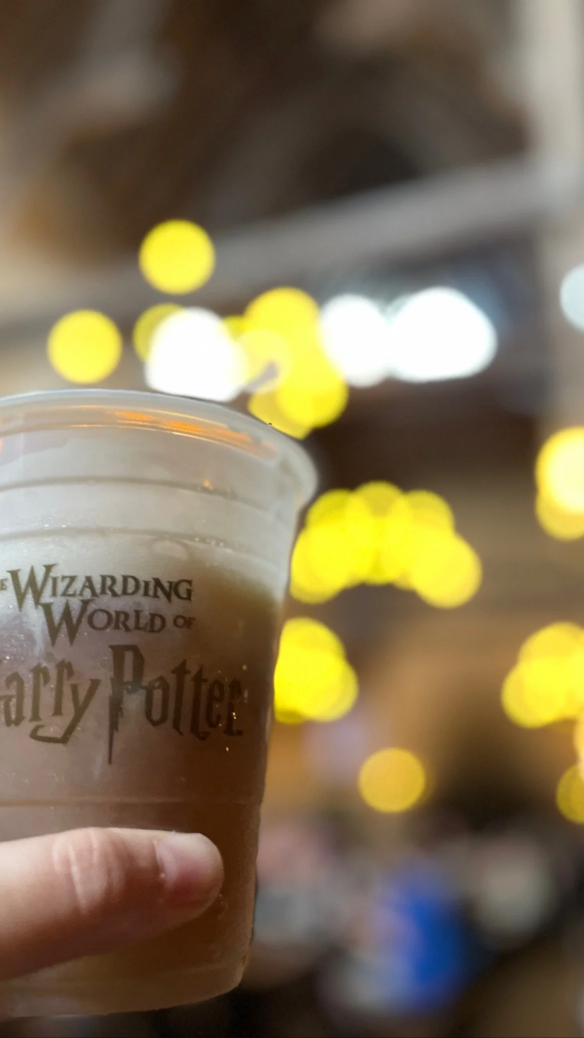 Close-up of a hand holding a plastic cup with the Harry Potter logo, against a background of yellow bokeh lights and blurred lights.