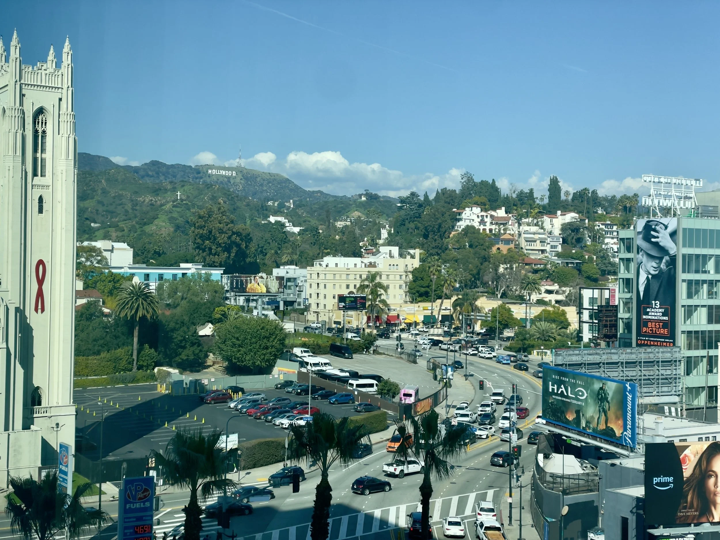 Cityscape of Hollywood, Los Angeles, showing the Hollywood sign on the hillside, commercial buildings, billboards, a busy street with cars, and palm trees under a partly cloudy sky.