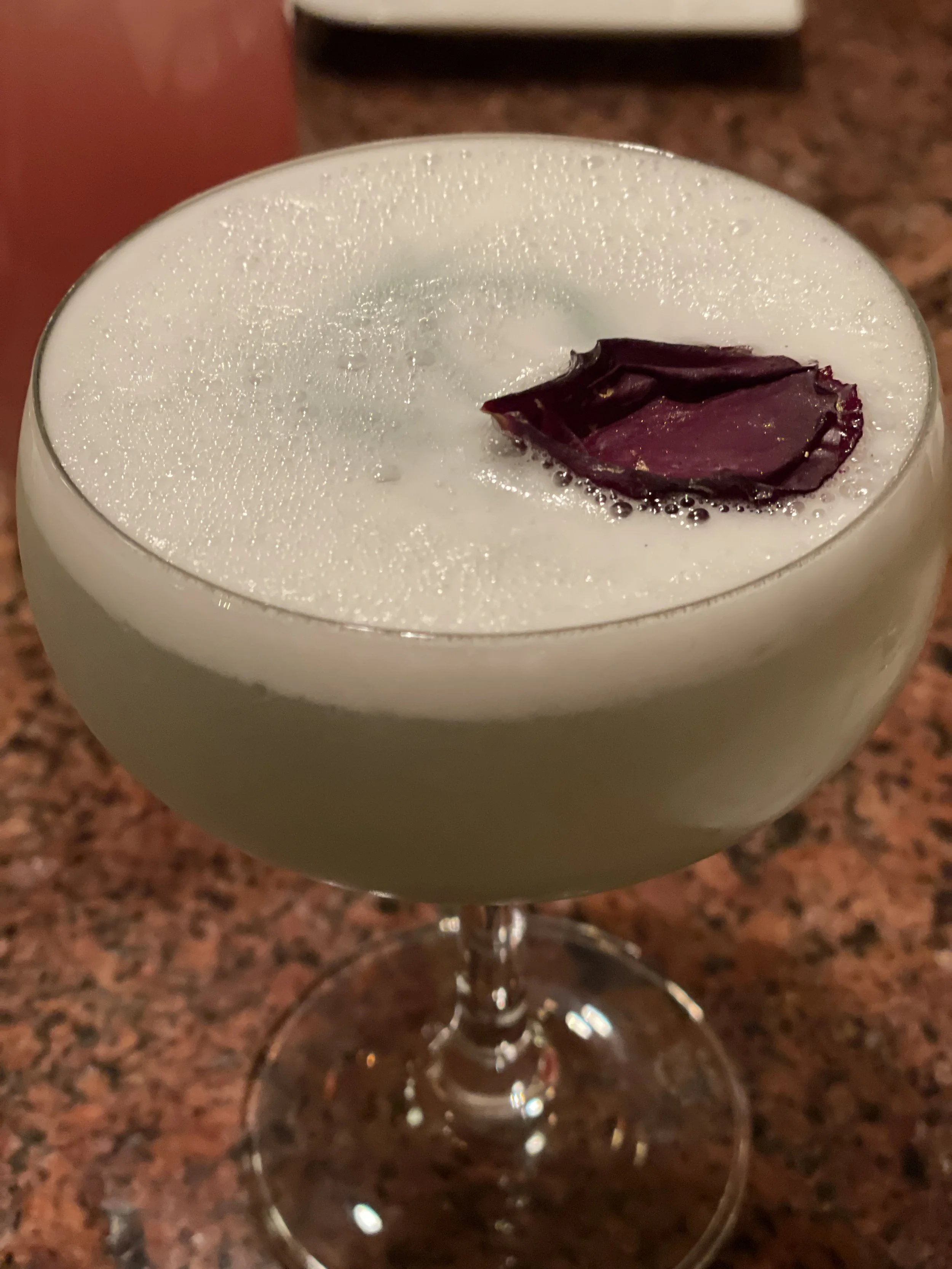 Cocktail in a coupe glass with foam and a purple flower petal garnish on top, placed on a granite countertop.