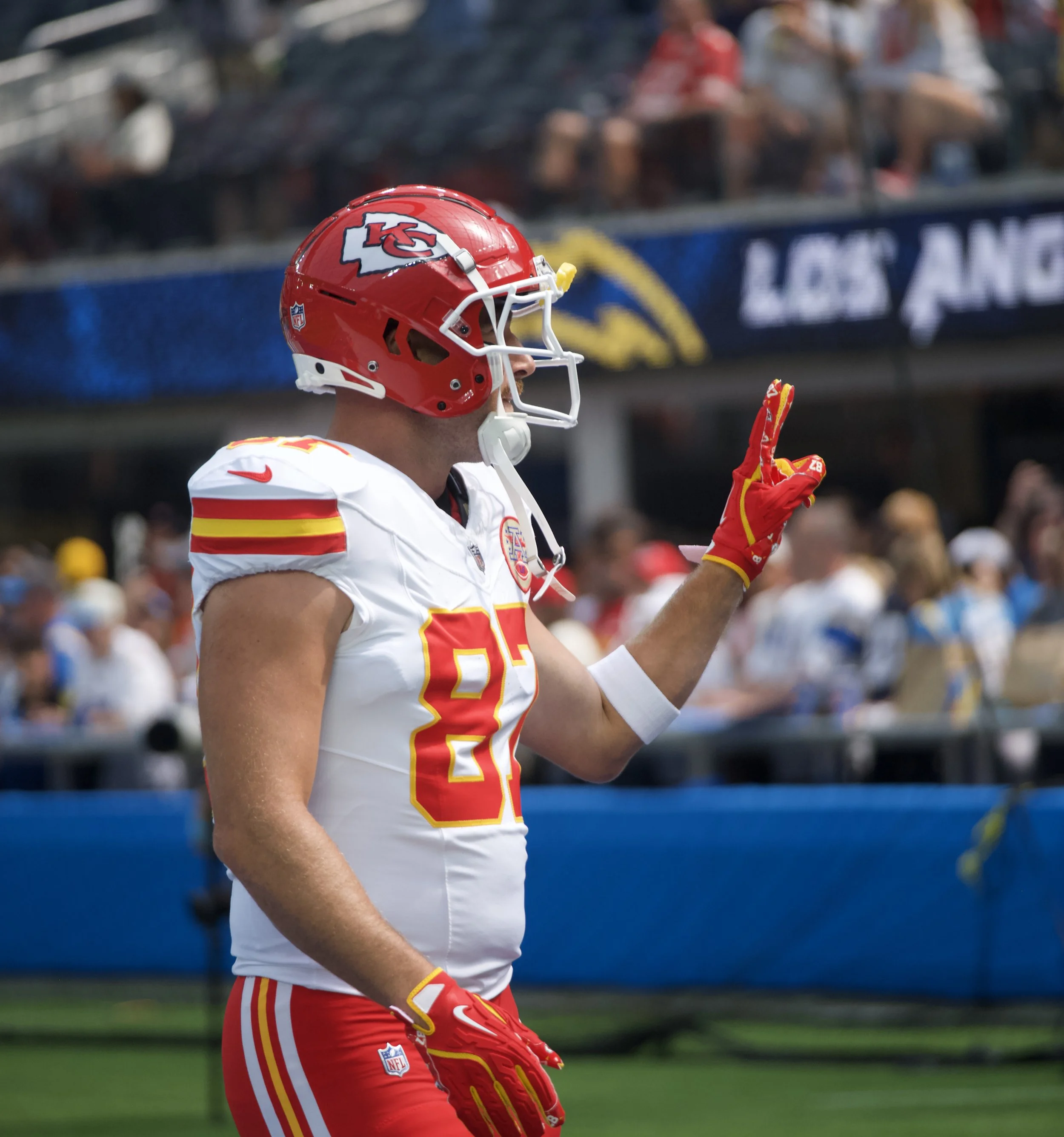 A football player in a red and white Kansas City Chiefs uniform on the field during a game, making a gesture with his right hand.