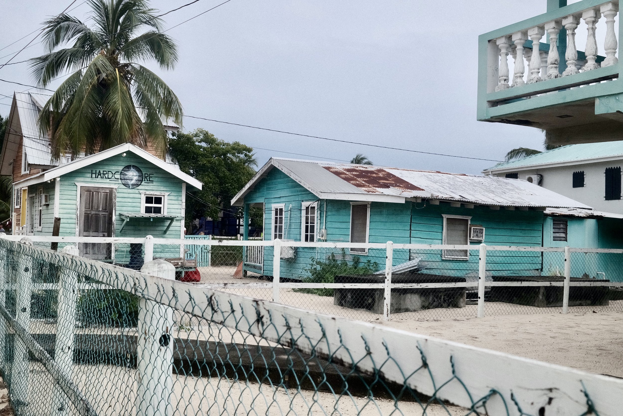 Two small turquoise houses with white trim and corrugated metal roofs, one with a rusty section, surrounded by a chain-link fence and set on sandy ground, with palm trees and overcast sky in the background.