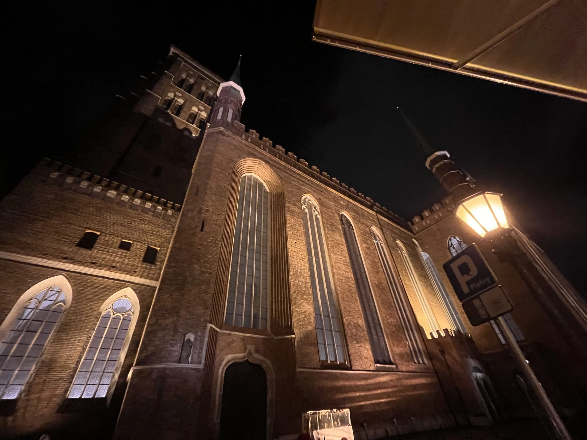 Nighttime view of a large, historical brick church with tall, arched windows illuminated from within and exterior lighting highlighting the structure. A street lamp and parking sign are visible in the foreground.