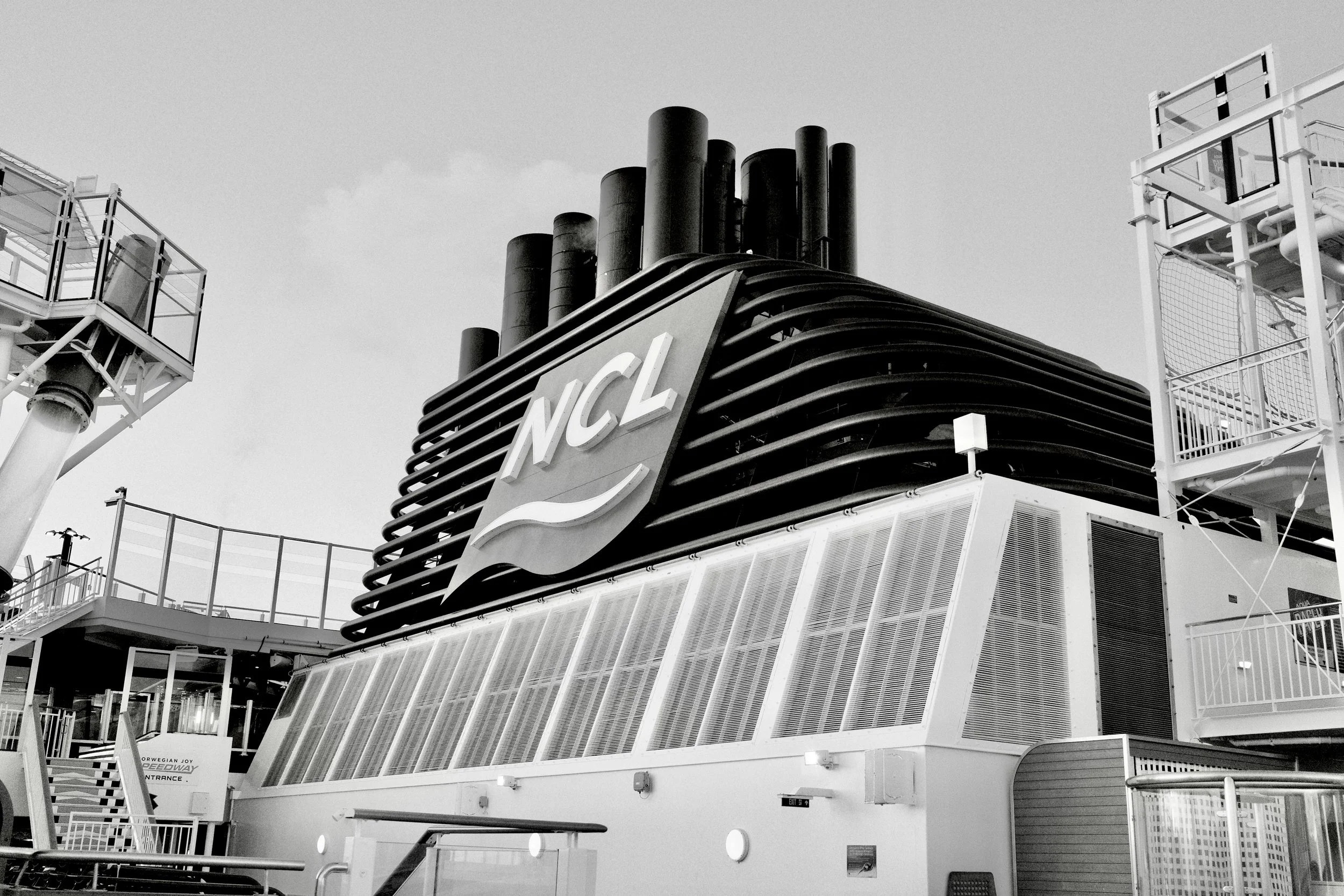 Black and white photo of a cruise ship's smokestack with the logo 'NCL' on it, surrounded by safety railings and part of the ship's deck structures.