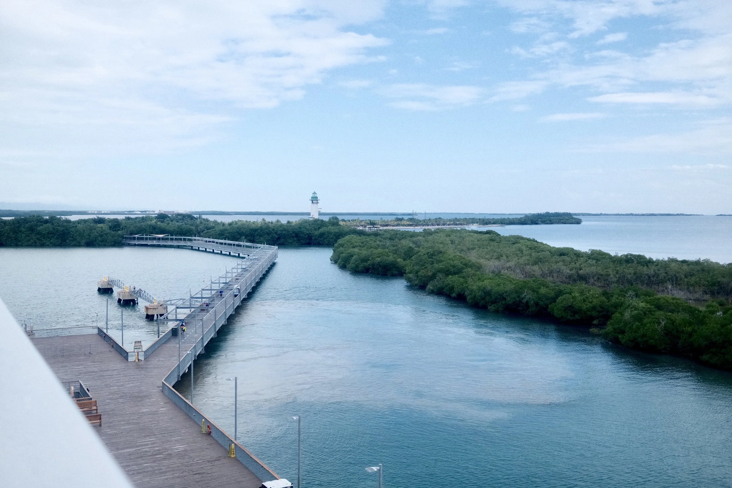 View of a pier extending into water with a lighthouse in the background, surrounded by trees and a partly cloudy sky.