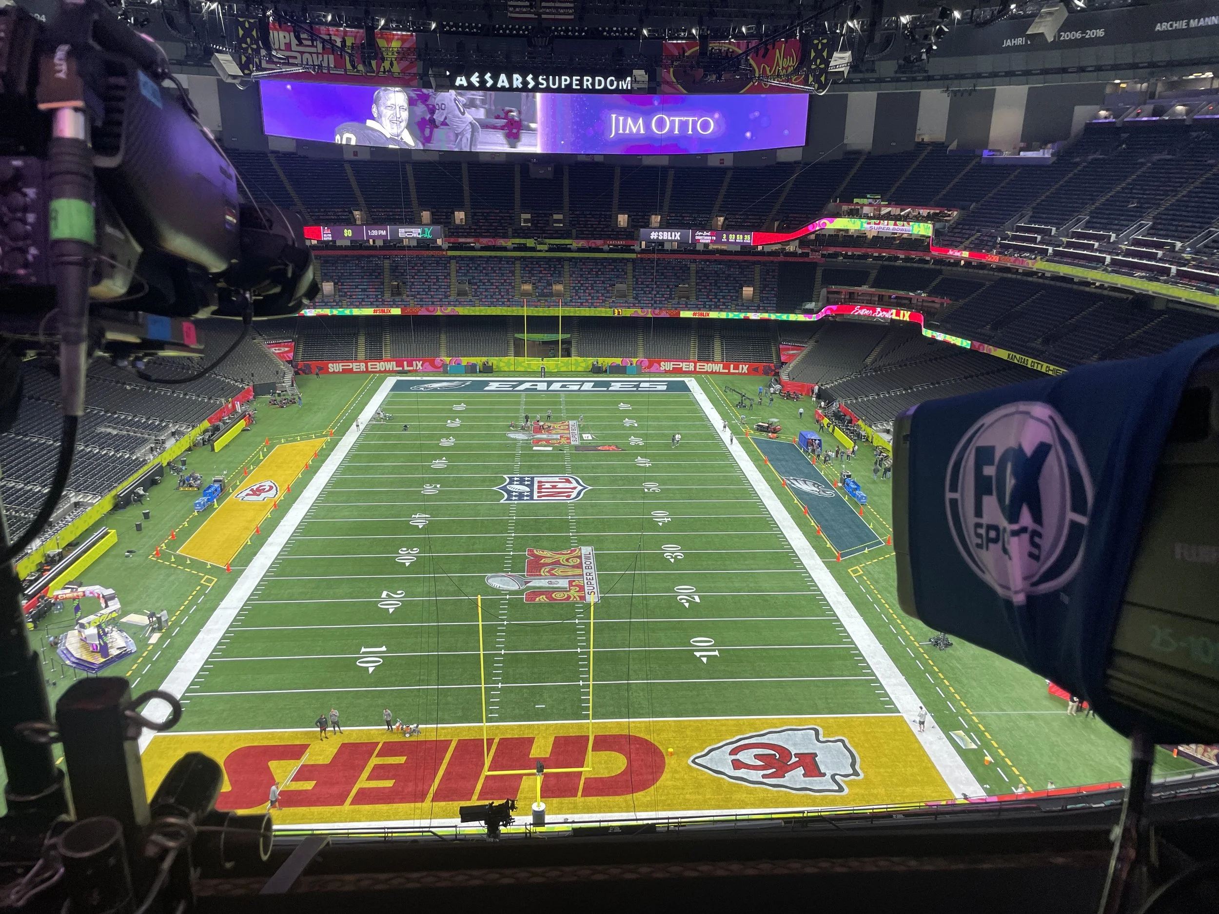 View from the 2025 Super Bowl, showing the field decorated Kansas City Chiefs' logo at the near end zone and the Philadelphia Eagles' logo at the far end zone. There are cameras and equipment in the foreground.