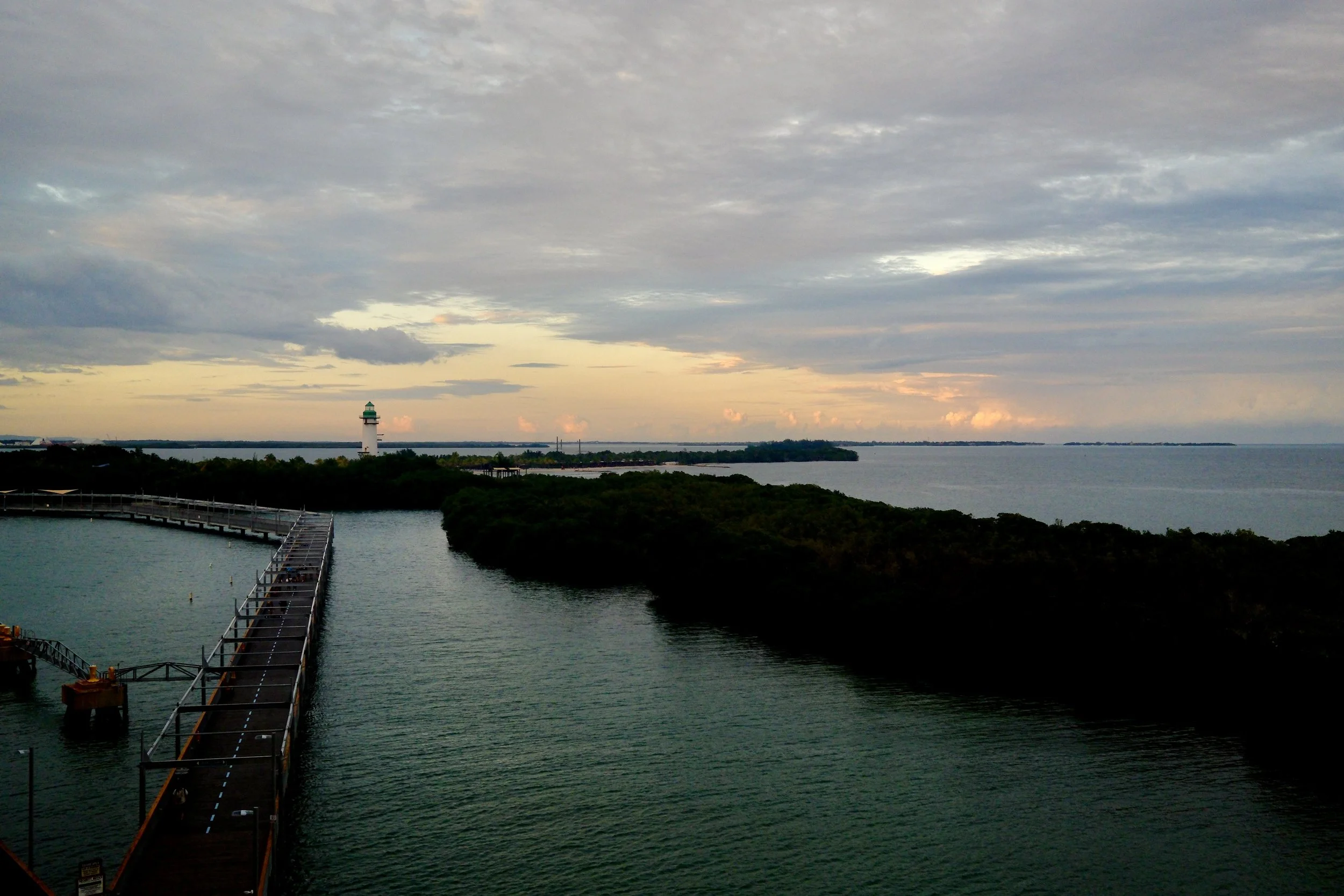 A scenic view of a lighthouse on a small island near a pier at sunset with cloudy sky and water surrounding the island.