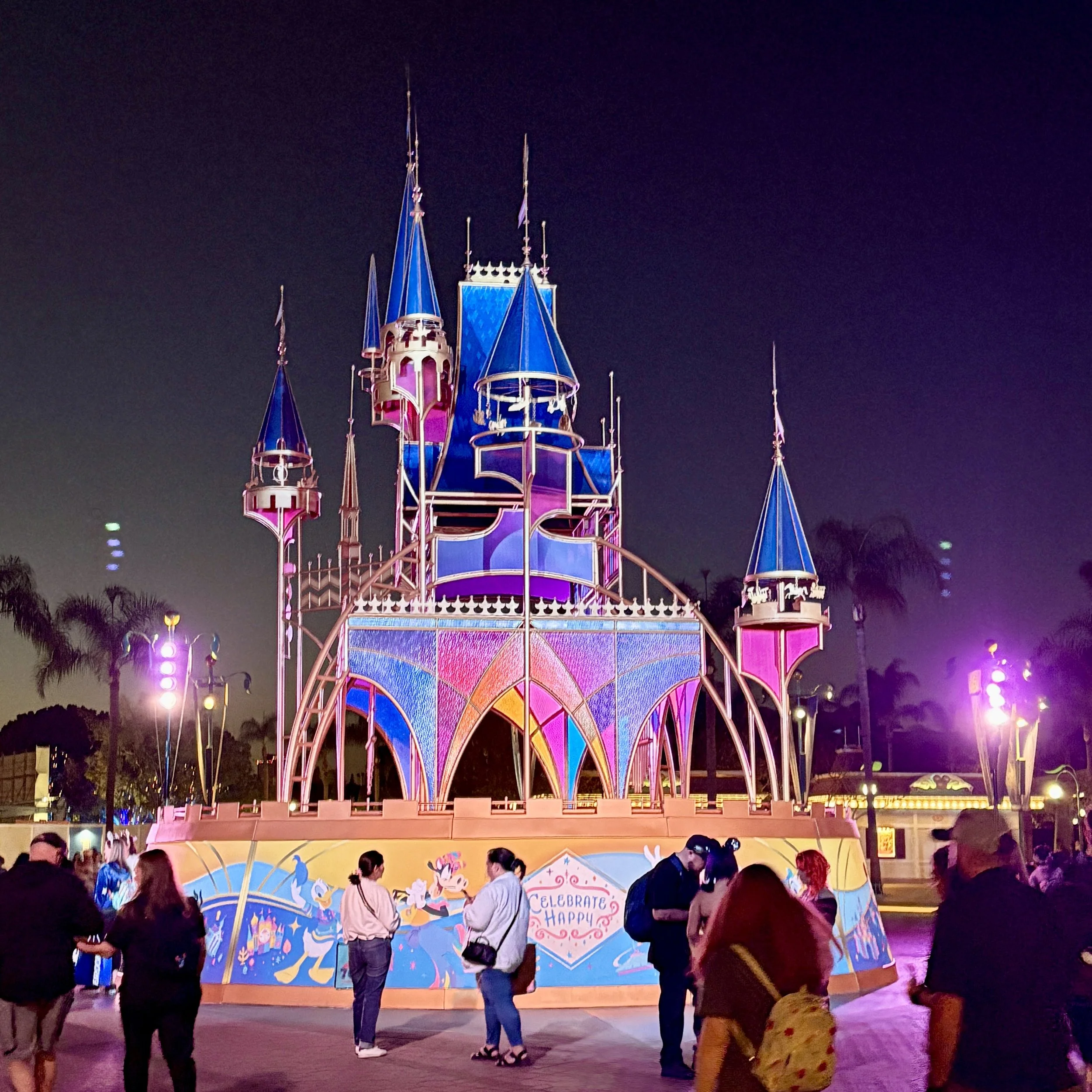 A colorful Disney castle illuminated at night with people gathered around in front.