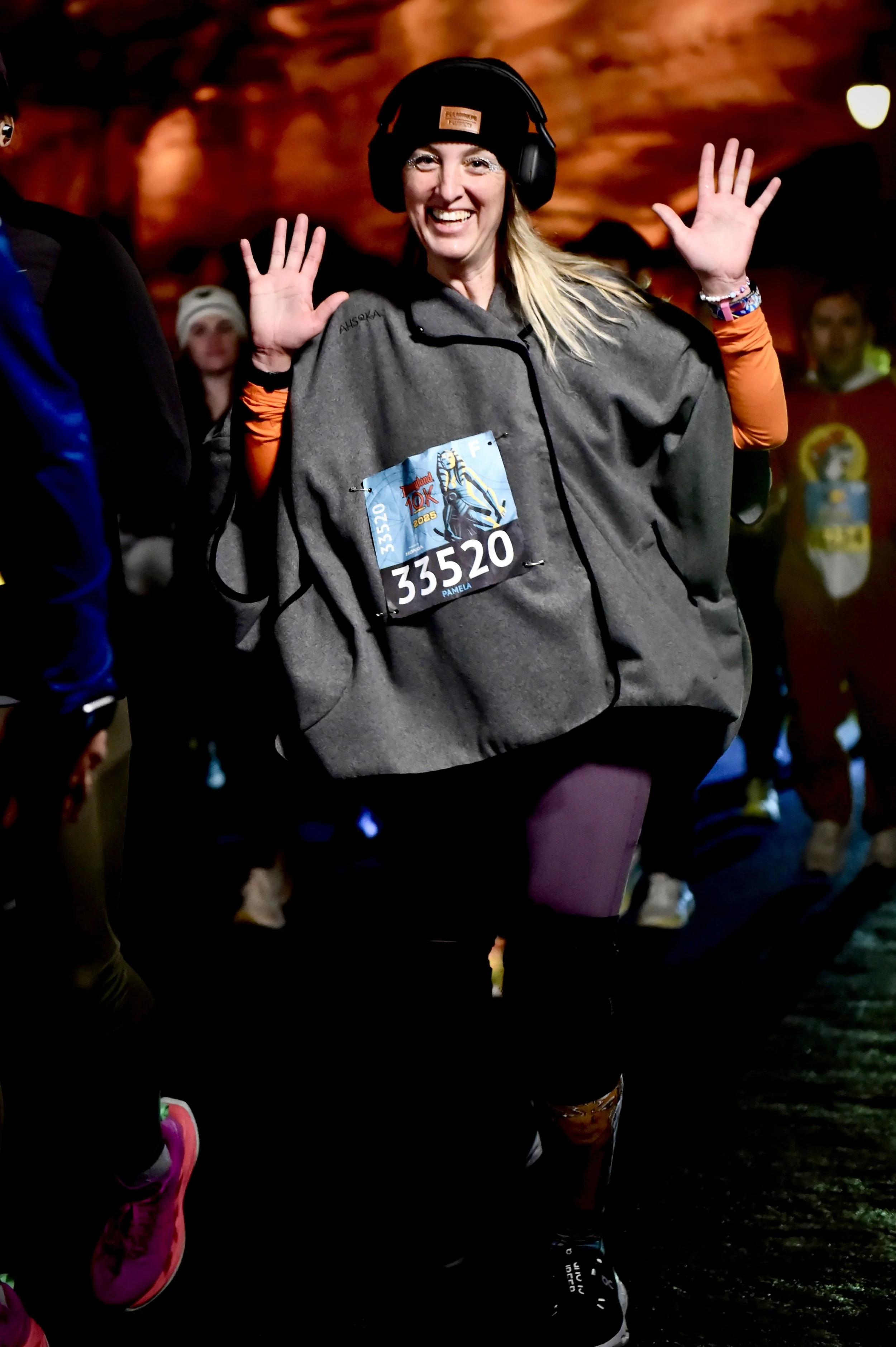 Pamela Bloom smiling and waving at a marathon event, wearing a beanie, headphones, a grey jacket, and a race bib at a runDisney event