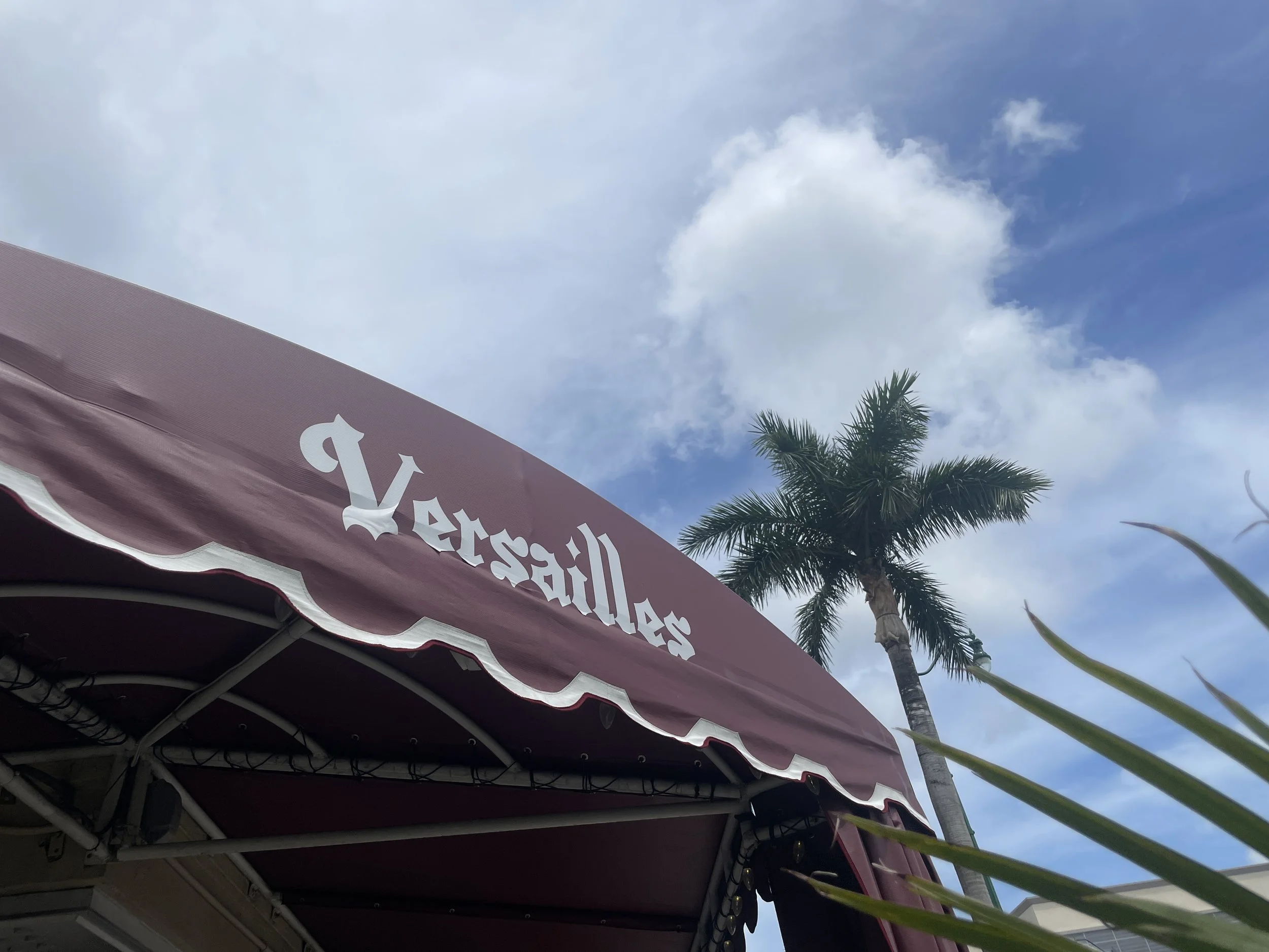 A maroon awning with the words "Versalles" in white Gothic font, a palm tree, blue sky with clouds, and some greenery in the foreground.