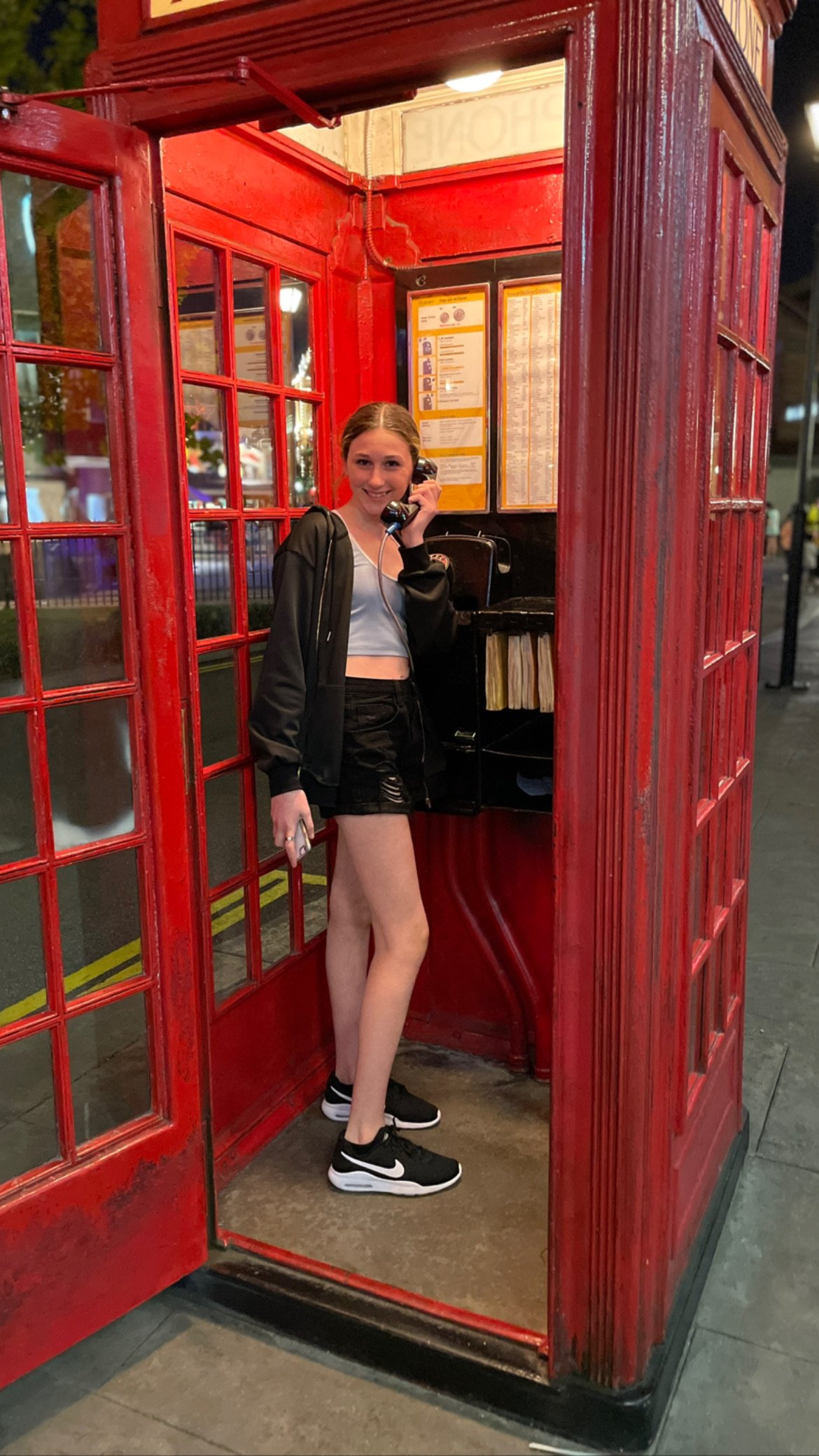 Young woman standing inside a classic red British telephone booth, talking on the phone and holding a mobile phone, at night