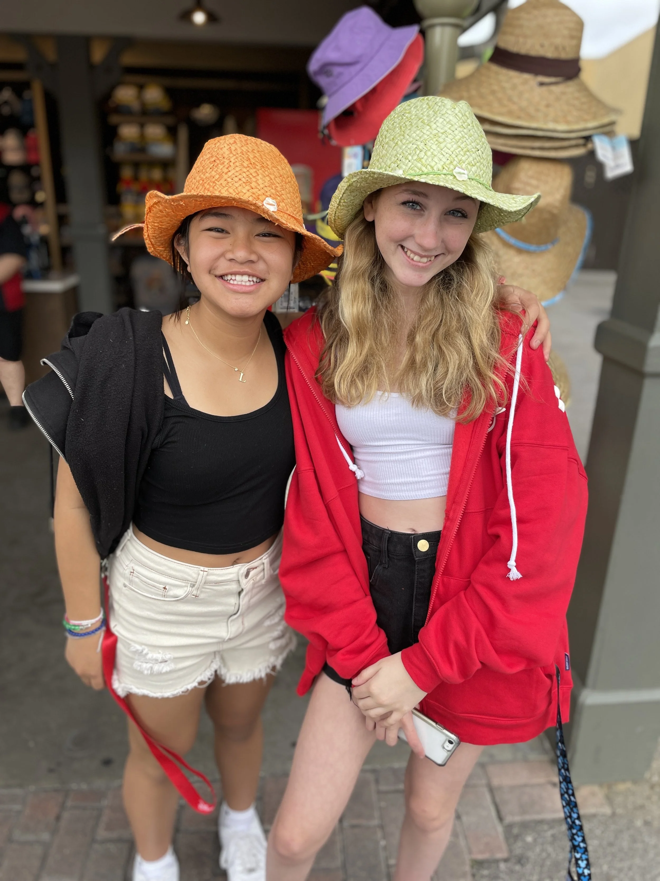 Two young women, one Asian and one Caucasian, wearing colorful straw hats, smiling, standing outside in front of a hat display shop.