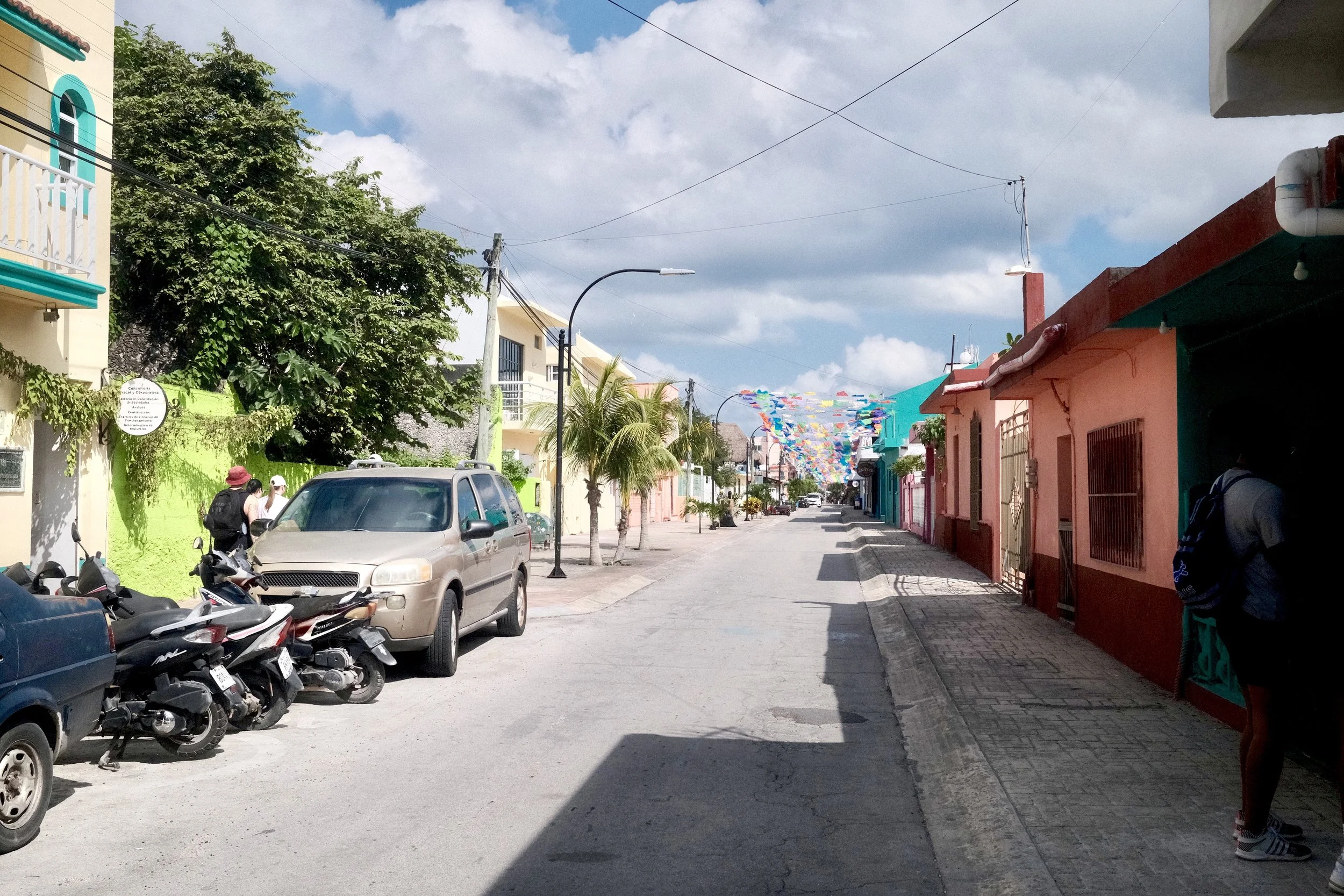 Colorful street with parked cars and motorcycles, palm trees, and buildings painted in bright colors under a partly cloudy sky, with festive banners strung across the street.