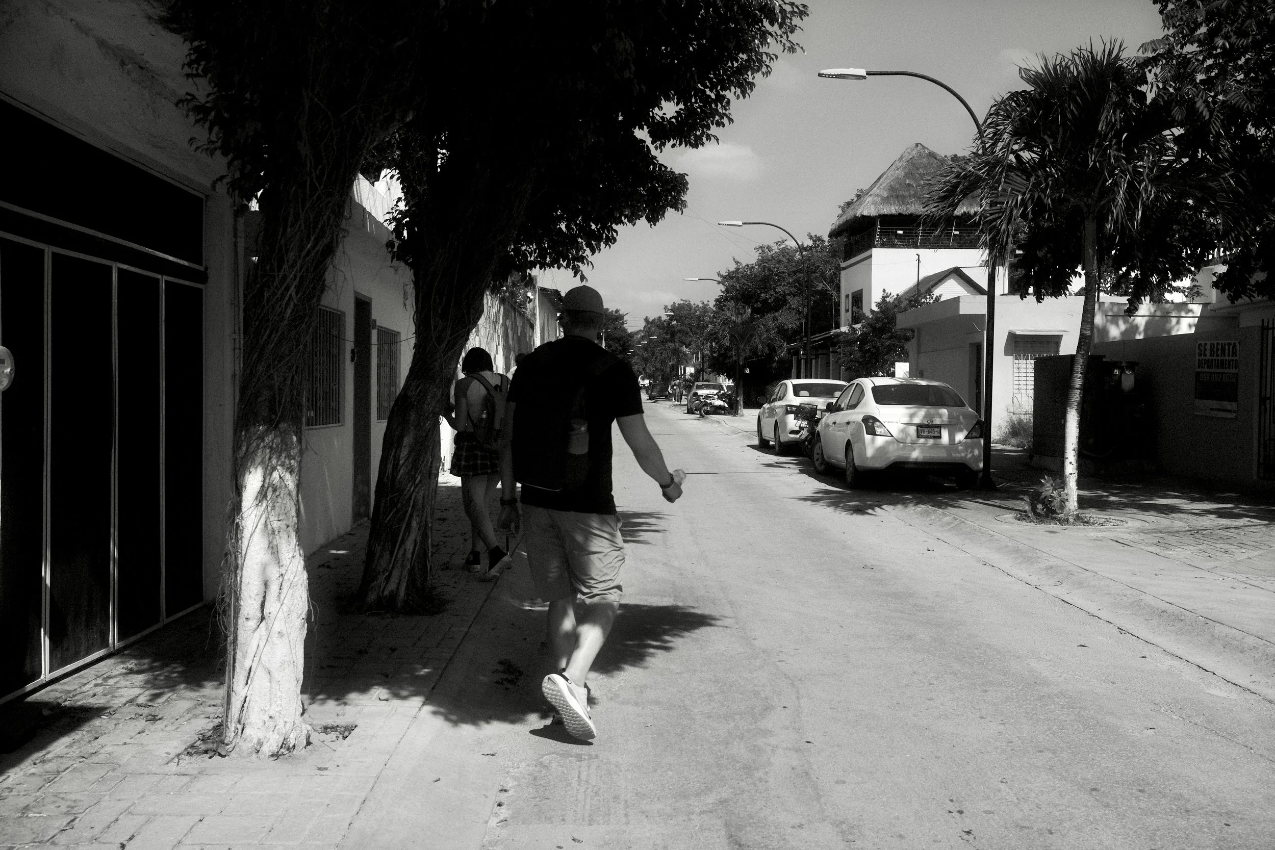 A person walking on a sidewalk in a neighborhood street with trees, parked cars, and houses in the background.