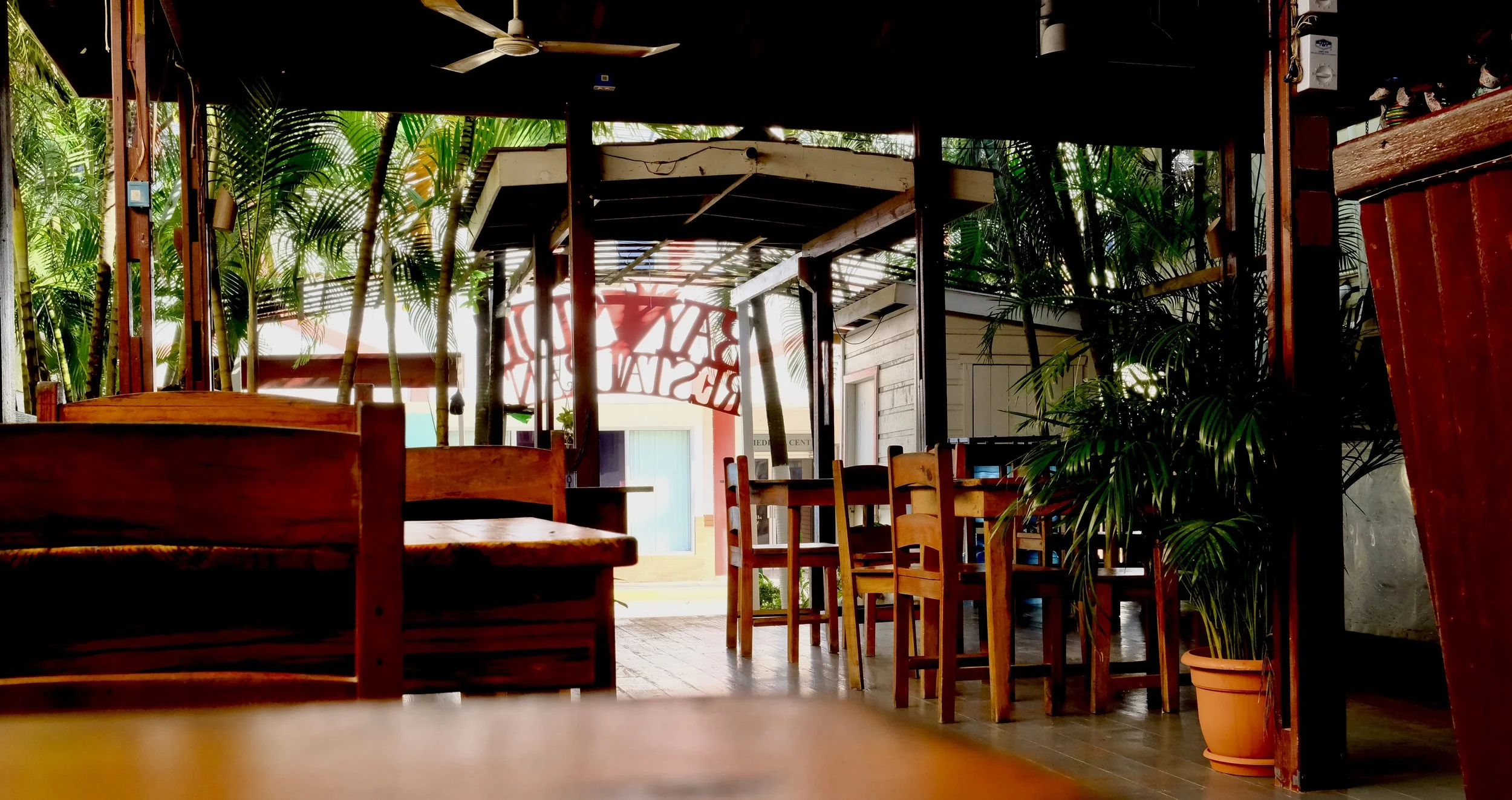 Interior of a tropical-themed restaurant with wooden furniture, potted plants, and greenery outside, seen through large windows.