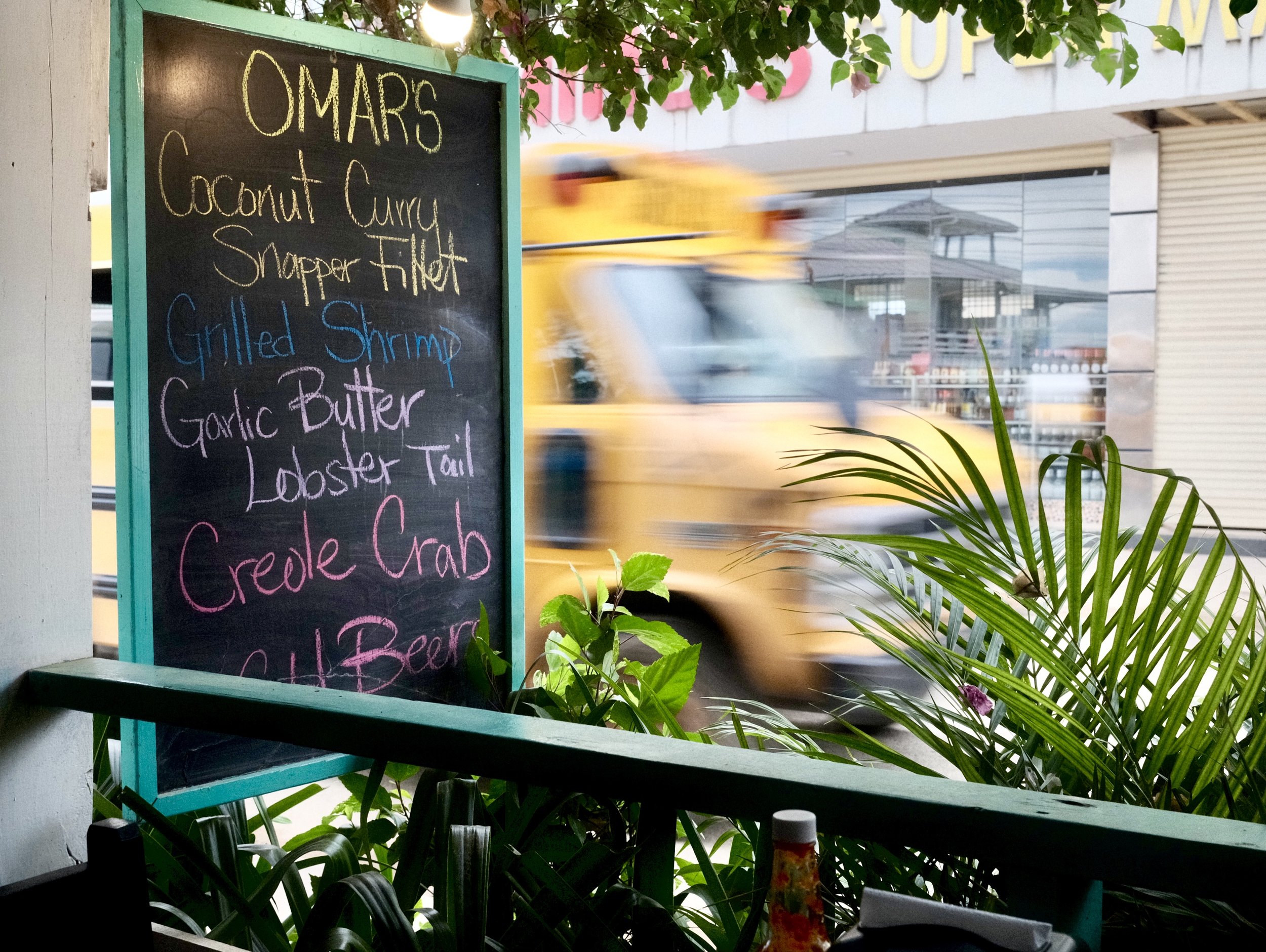 A colorful chalkboard sign listing seafood items outside a restaurant, with a blurred yellow taxi passing by and tropical plants in the foreground.