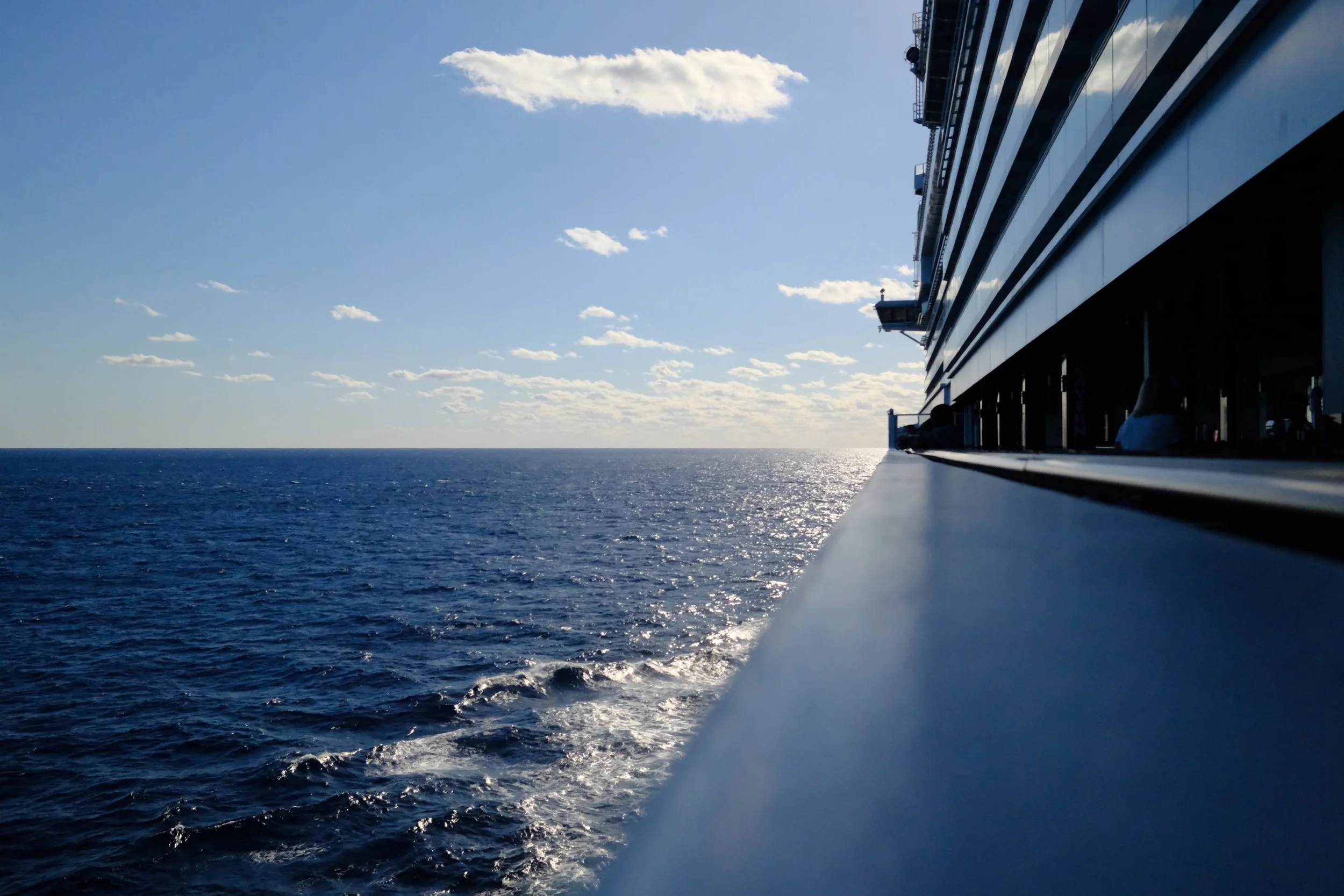 View of the open ocean from a cruise ship balcony during daytime, with a clear blue sky and some scattered clouds.