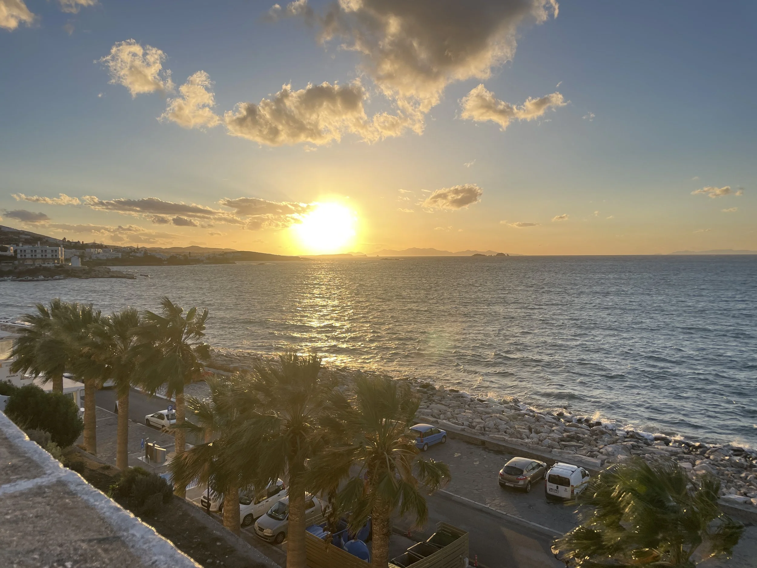 Sunset over the ocean with a partly cloudy sky, palm trees, cars parked along a coastal road, and buildings in the distance.