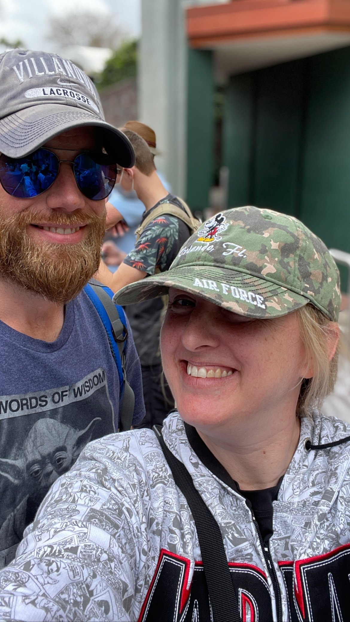 A man with a beard wearing sunglasses, a Villanova lacrosse cap, and a blue T-shirt, smiling for a selfie with a woman wearing a camouflage "Air Force" cap and a white patterned hoodie, both smiling. Other people are in the background.