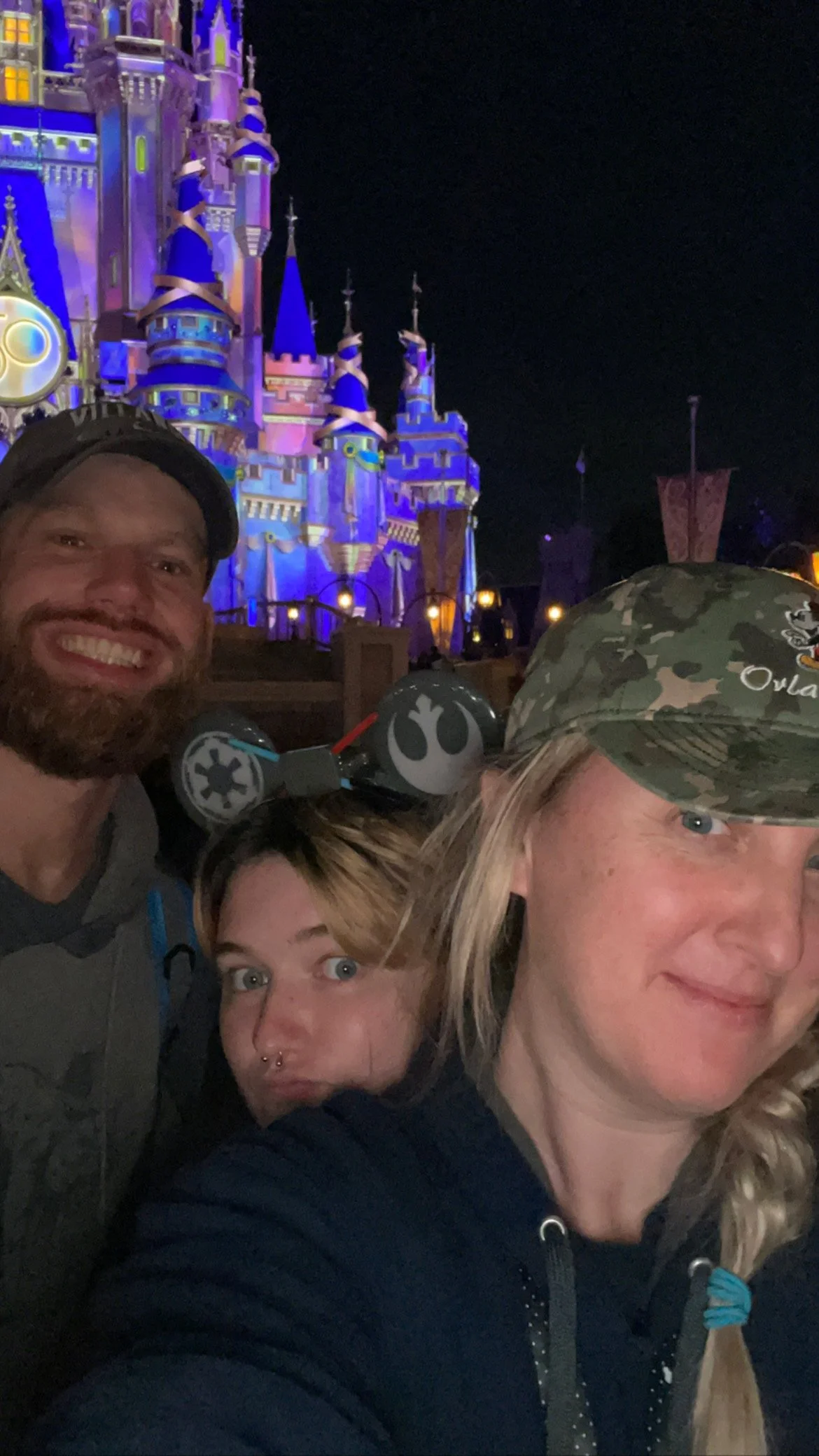 Three people taking a selfie in front of Sleeping Beauty Castle at Disneyland during the night, with the castle illuminated in purple and blue lights.