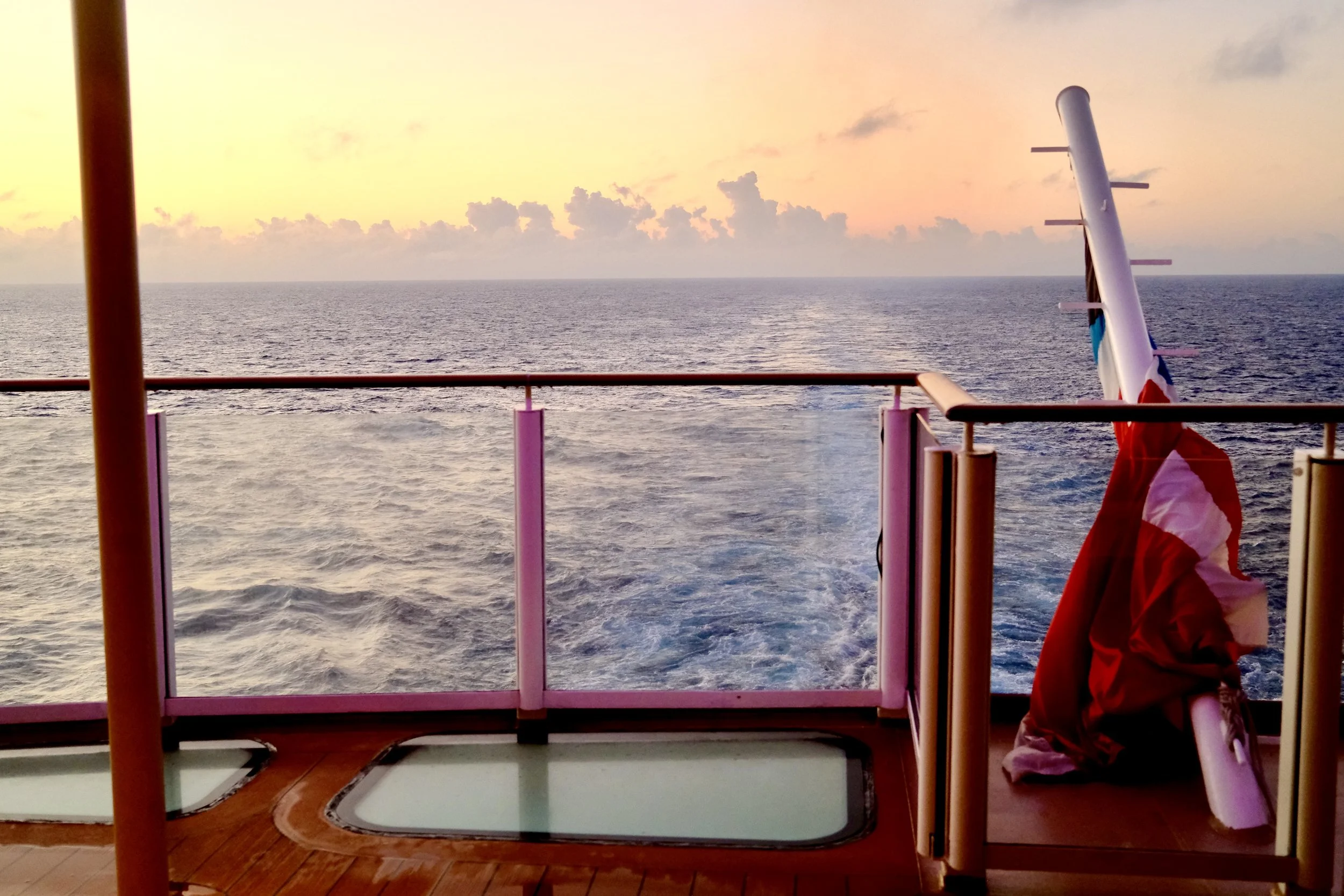 View of the ocean at sunset from a cruise ship balcony, with a French flag and a paddleboard leaning against the railing.