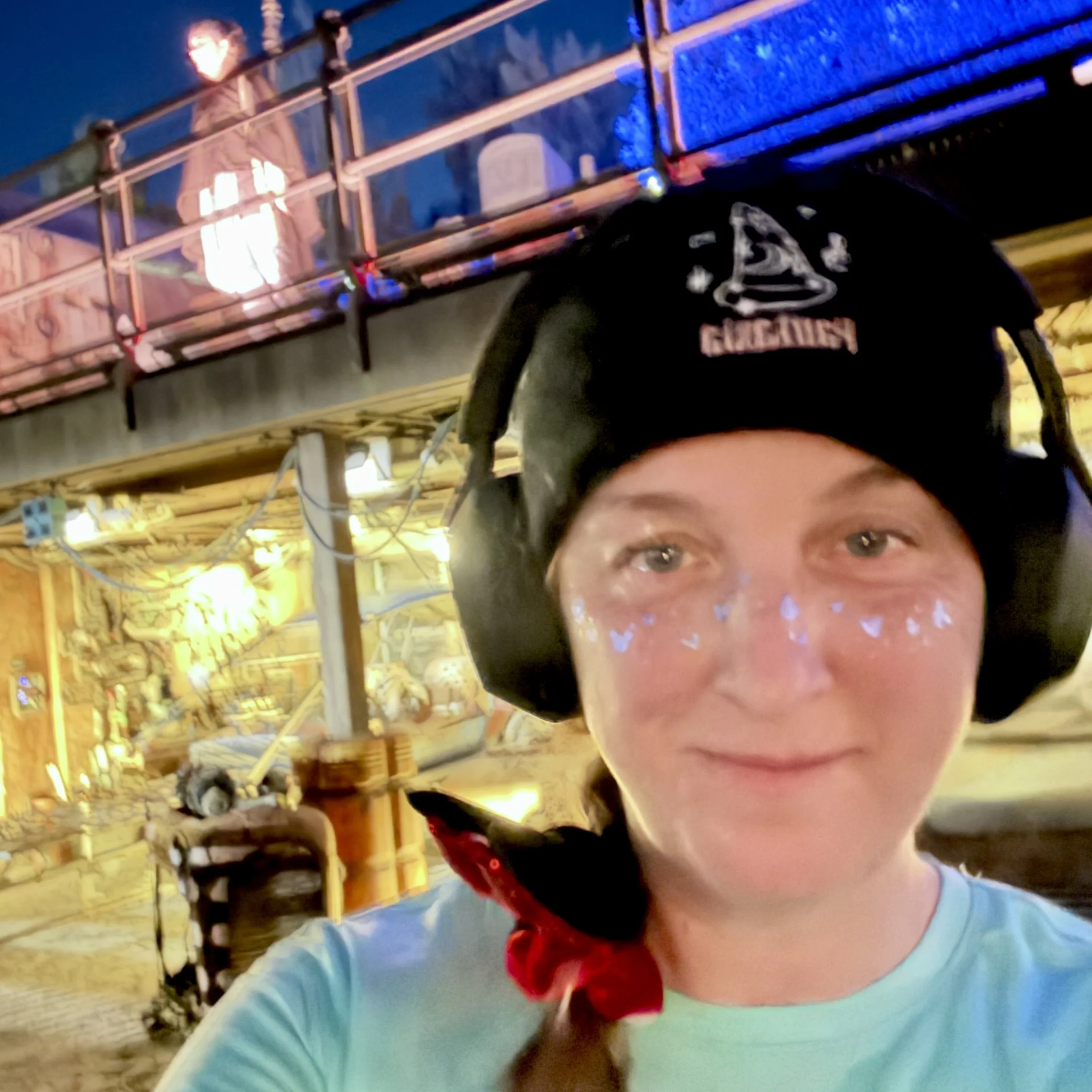 A young person wearing a black beanie with 'Glocklen' logo and large headphones, smiling at the camera with face paint dots. They are outdoors with a wooden structure and colorful lights in the background.
