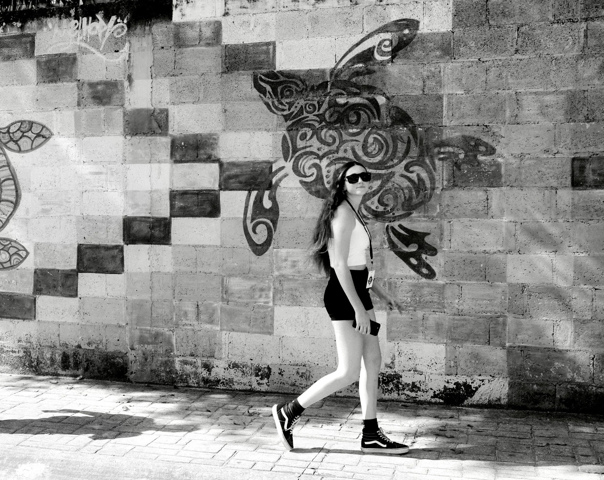 A young woman with long hair wearing sunglasses, a sleeveless top, shorts, and sneakers walking past a brick wall with a graffiti artwork of a butterfly.