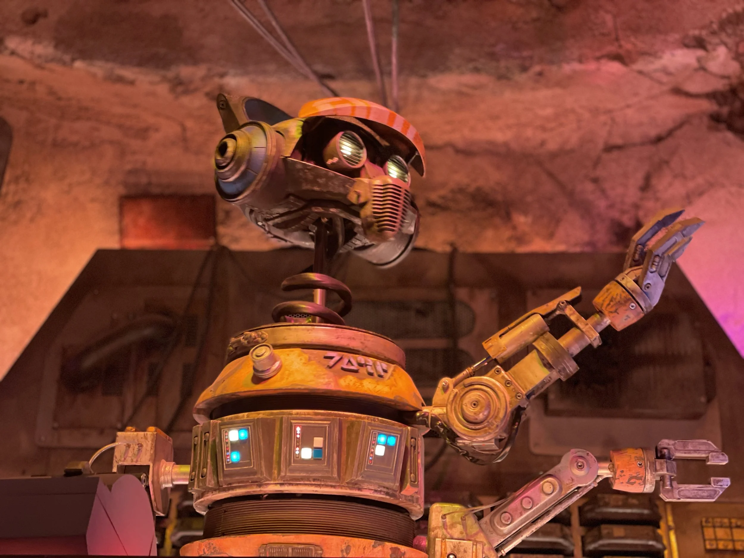 Close-up of a vintage robot with a round orange head, large circular eyes, and robotic arms, making a peace sign with one arm in a dimly lit setting with warm pink and purple lighting.