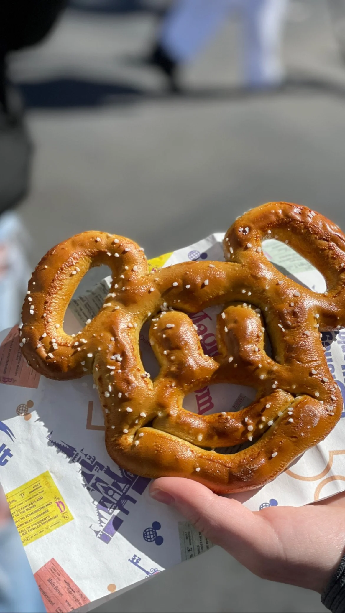 A large, golden-brown pretzel with coarse salt on top, being held by a person's hand over a paper bag at an outdoor location.