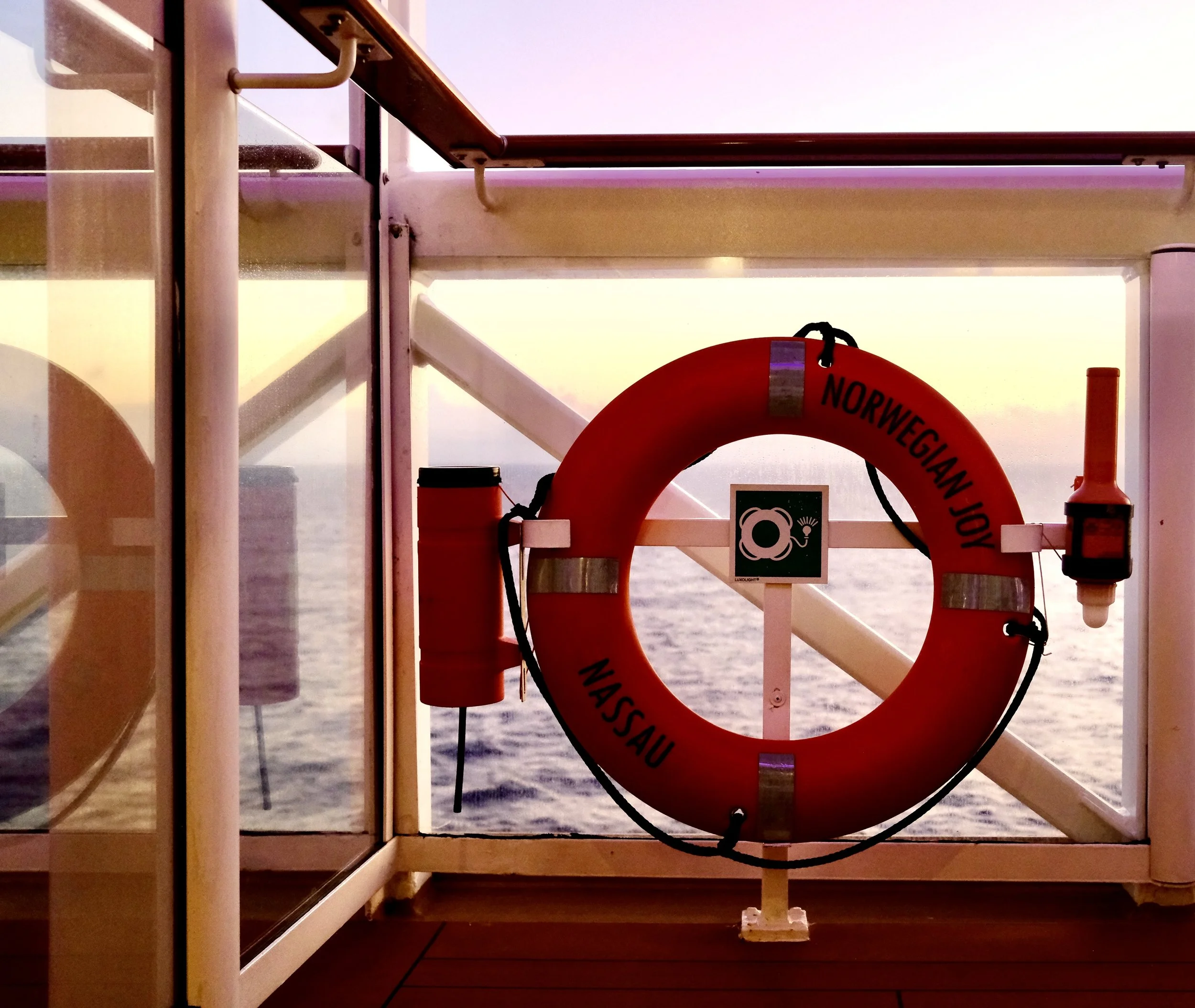 Life preserver ring with 'Norwegian Joy Nassau' written on it, hanging on a ship's side rail with a view of the ocean at sunset in the background.
