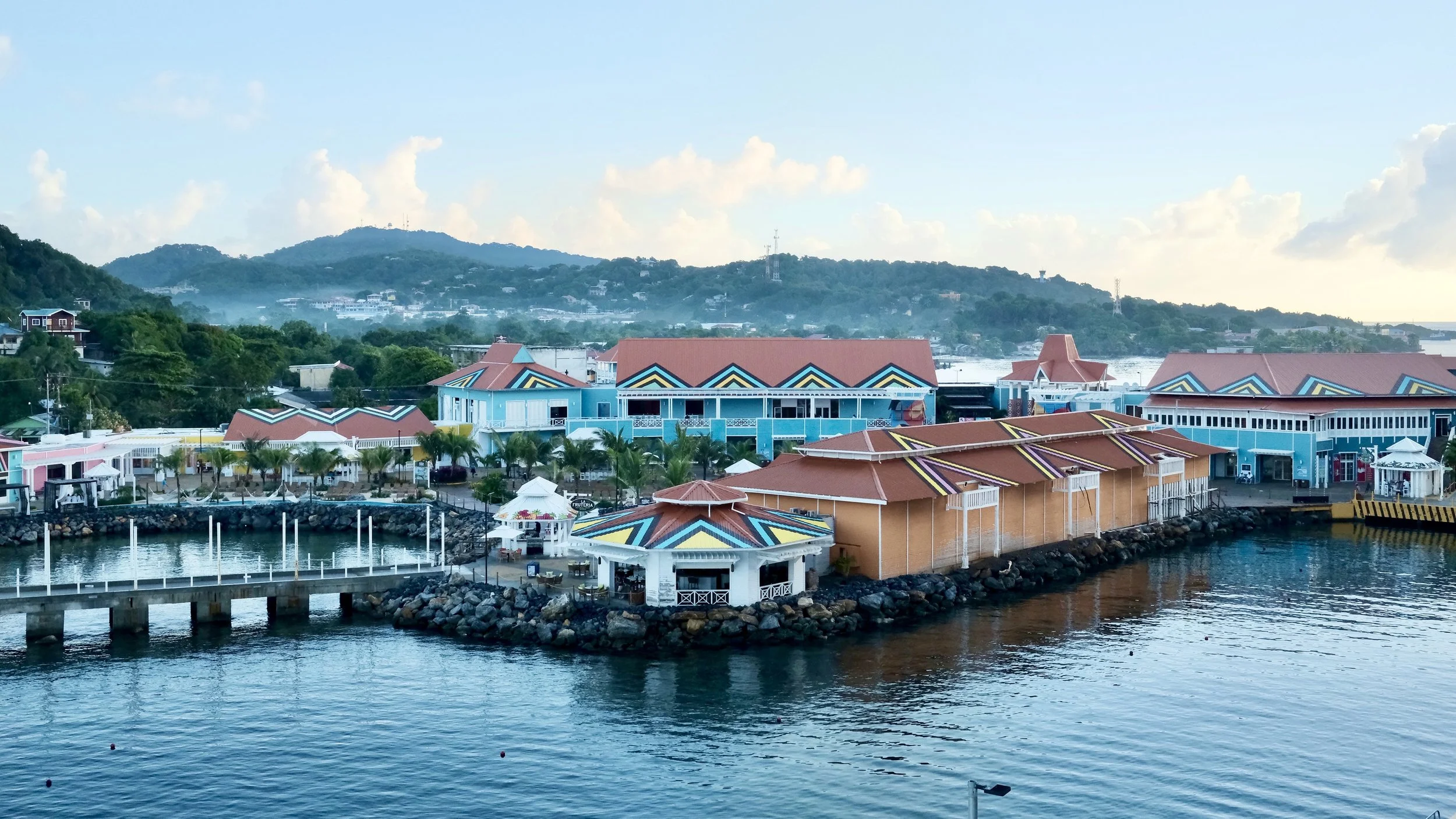 Colorful buildings with red, blue, yellow, and black roofs along a waterfront with water in the foreground and green hills in the background.