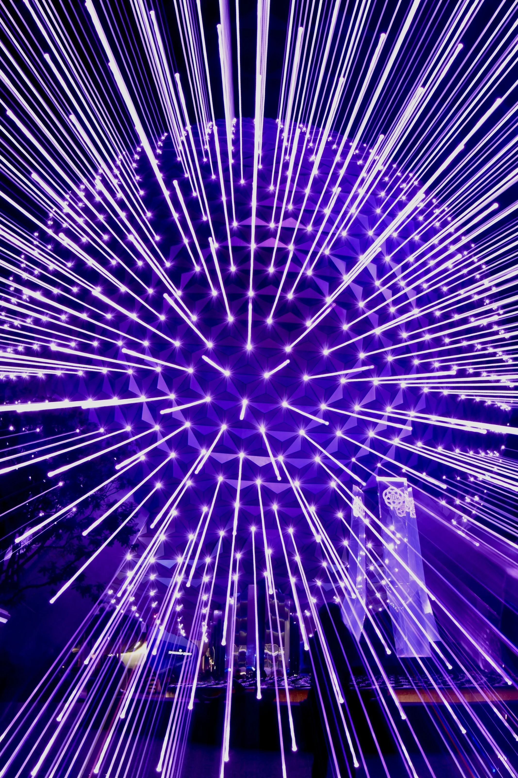 A display of vibrant purple and white light beams radiating outward in a spherical pattern against a dark background.