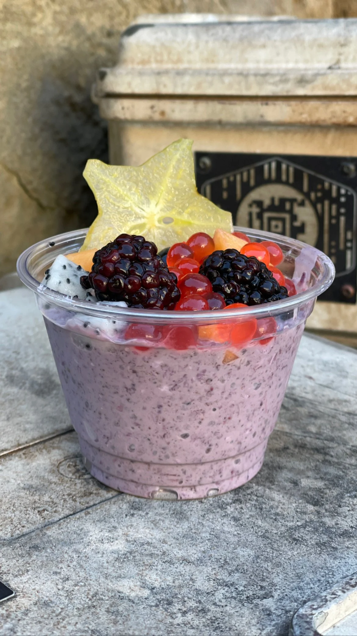 A clear plastic cup filled with a purple yogurt topped with various fresh fruits including blackberries, red currants, starfruit, and dragon fruit on a textured stone surface.