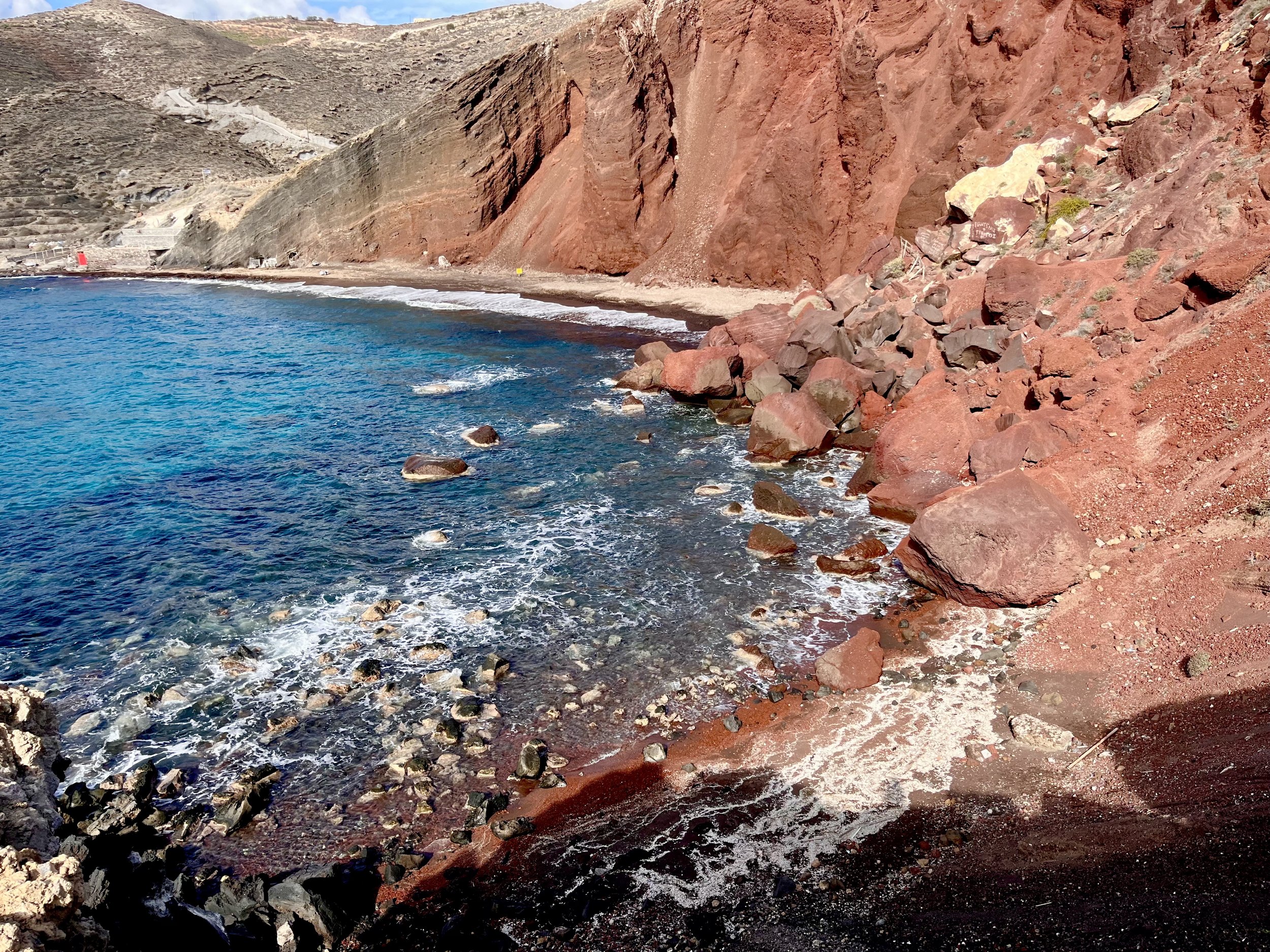 A rocky coastline with red cliffs adjacent to blue ocean water, with small waves, rocks, and a sandy shore.