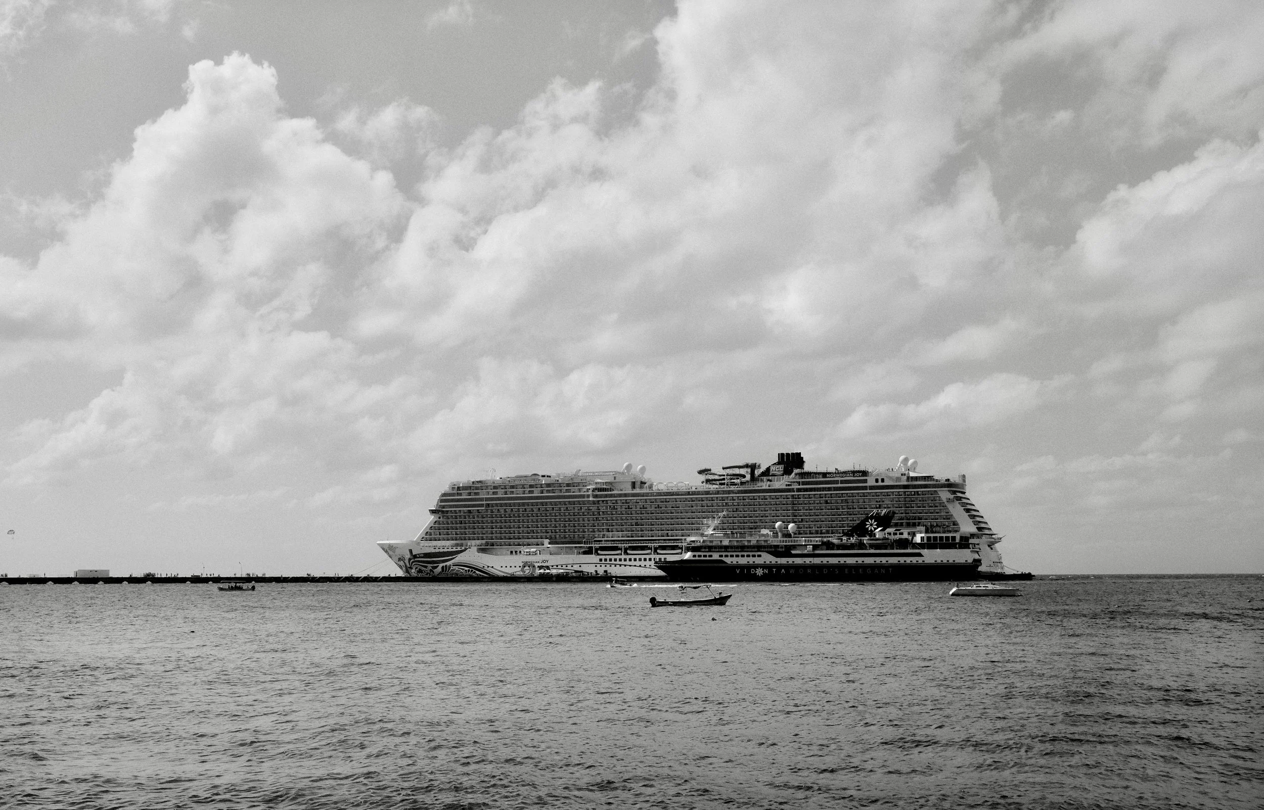 A large cruise ship sailing on the open water with a few small boats nearby, under a partly cloudy sky.