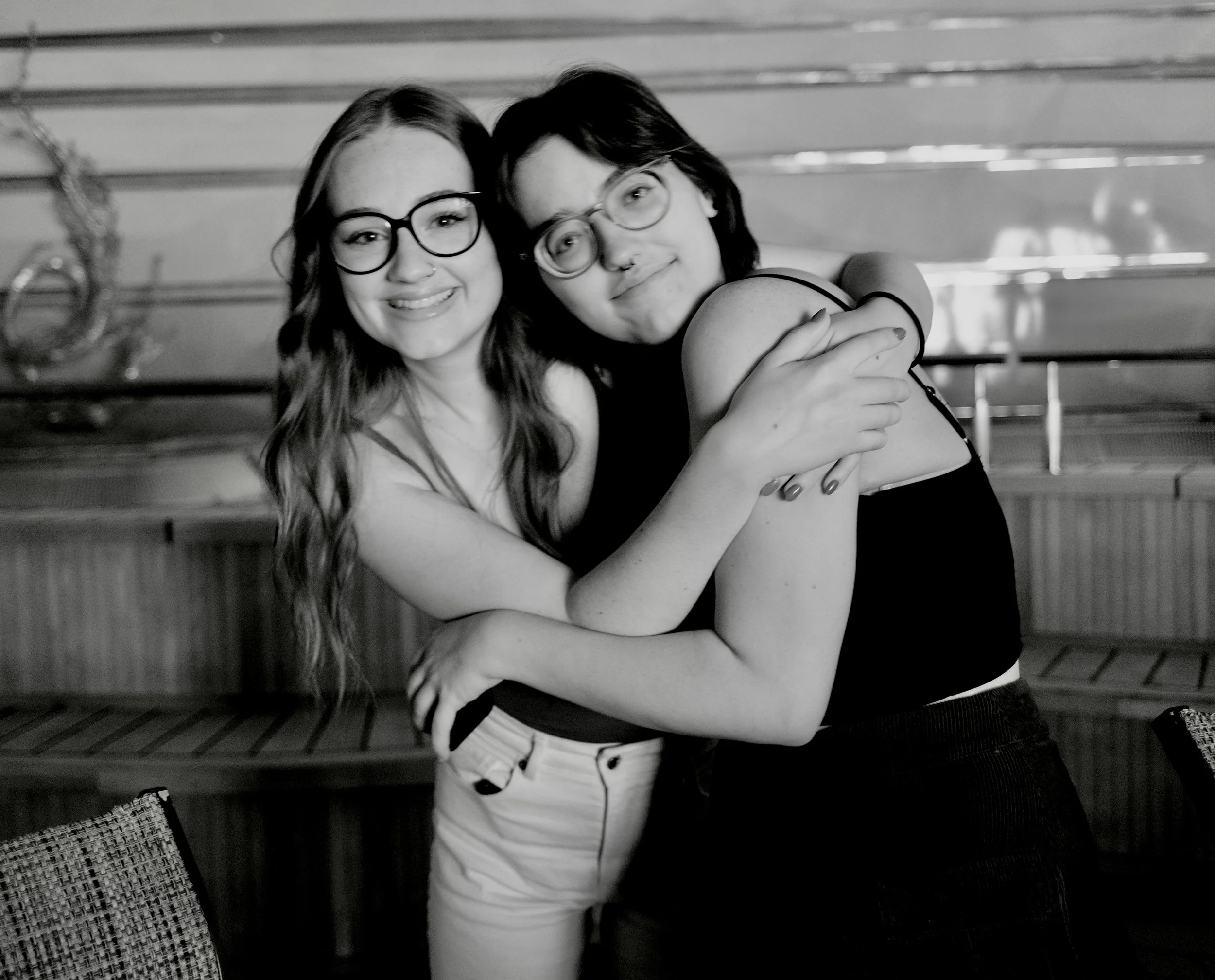 Two young women with glasses embracing and smiling. One has long wavy hair and the other has short dark hair. They are indoors with a wooden background.