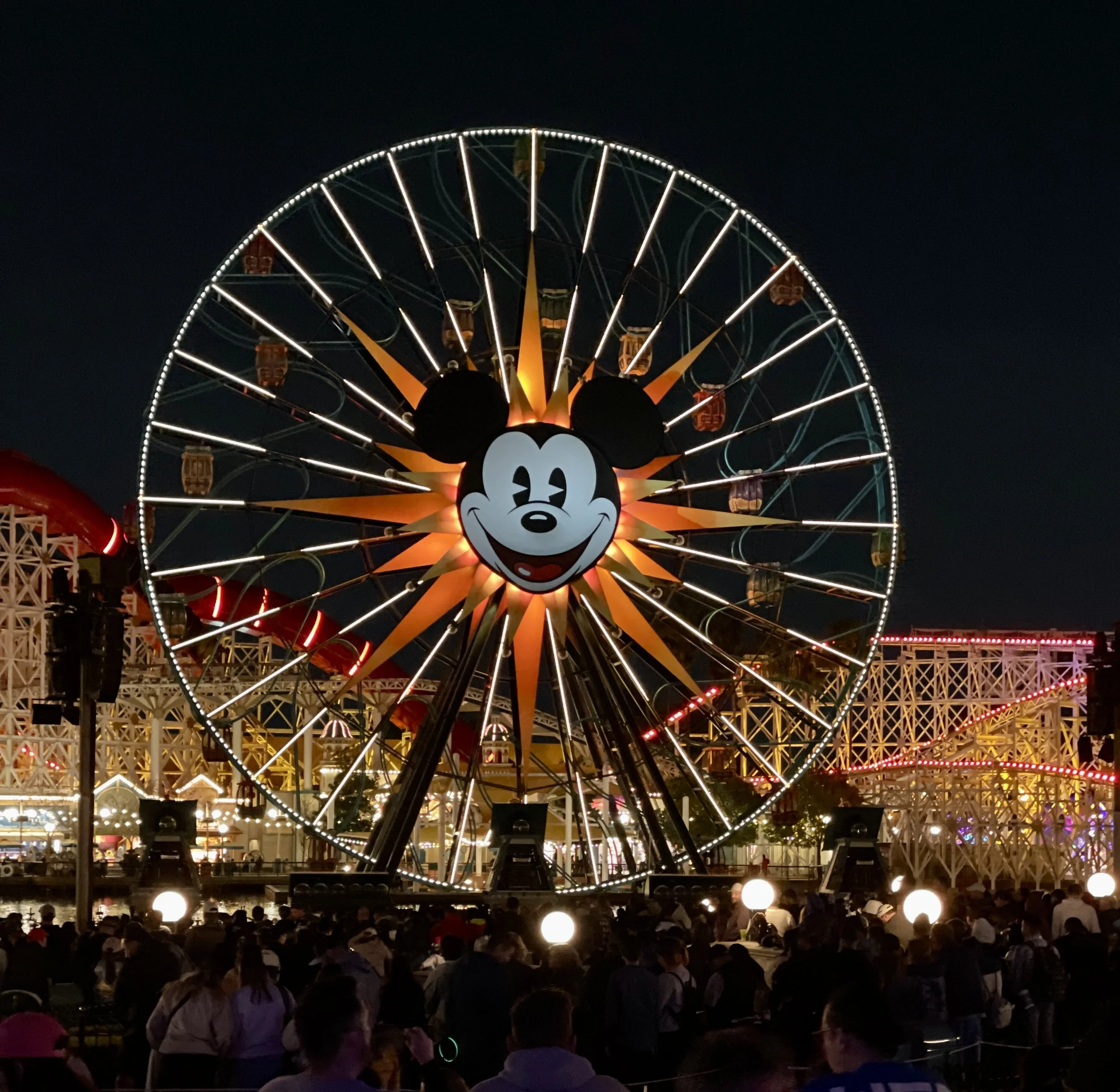 Night scene at a carnival with a Ferris wheel featuring Mickey Mouse's face at the center, surrounded by illuminated decor and a crowded audience.