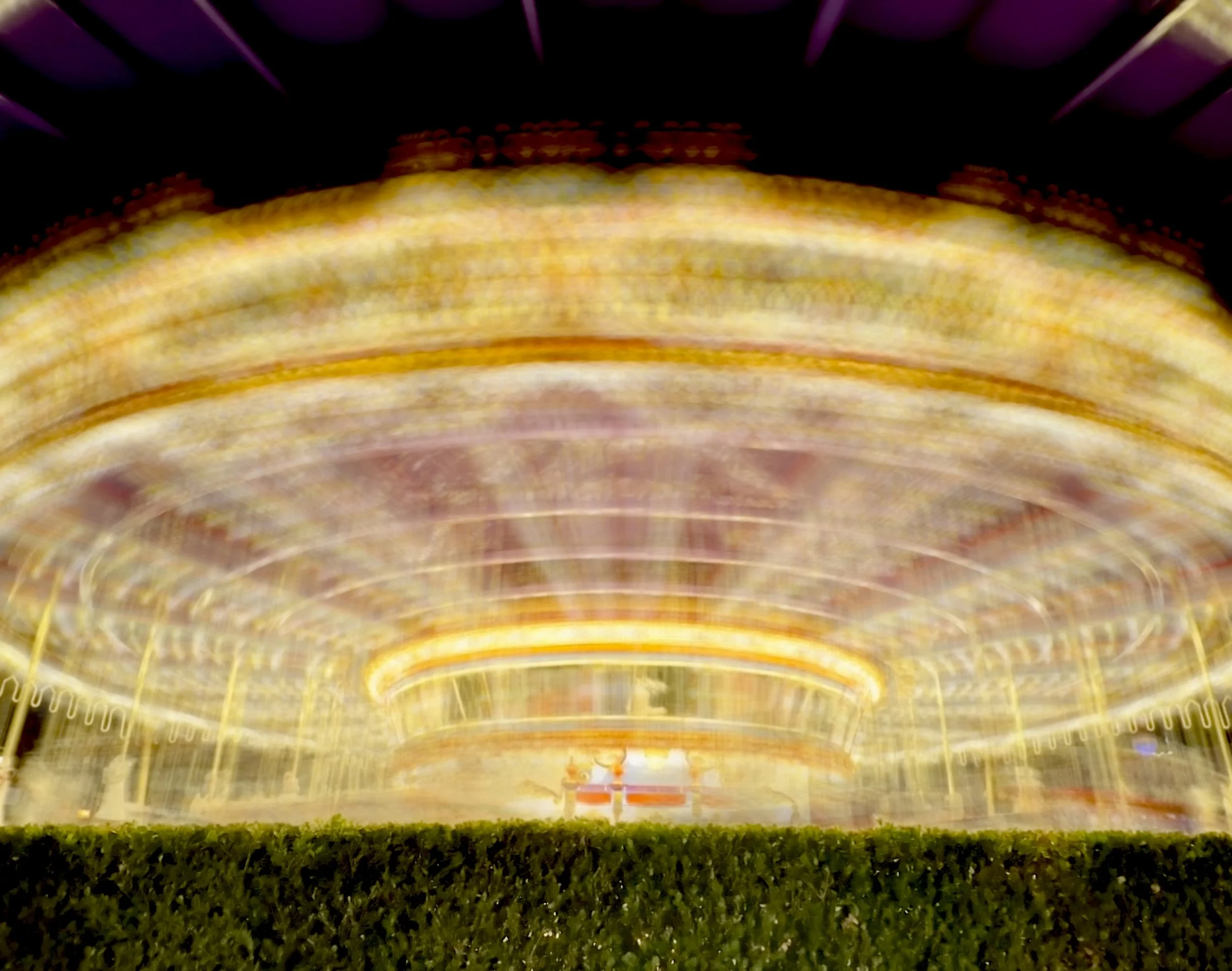 Long exposure photo of a brightly lit carousel at night, with blurred and swirling lights, and a green hedge in the foreground.